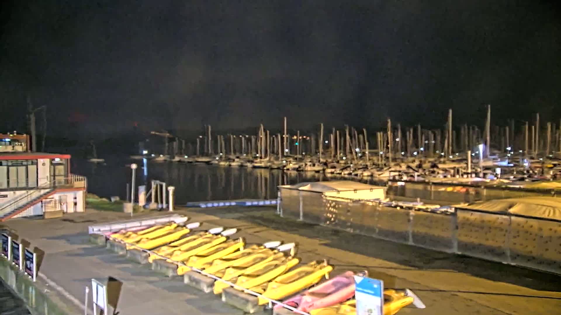 At night, a marina filled with docked sailboats is visible across a calm body of water from a waterfront area where numerous yellow kayaks are lined up for storage.