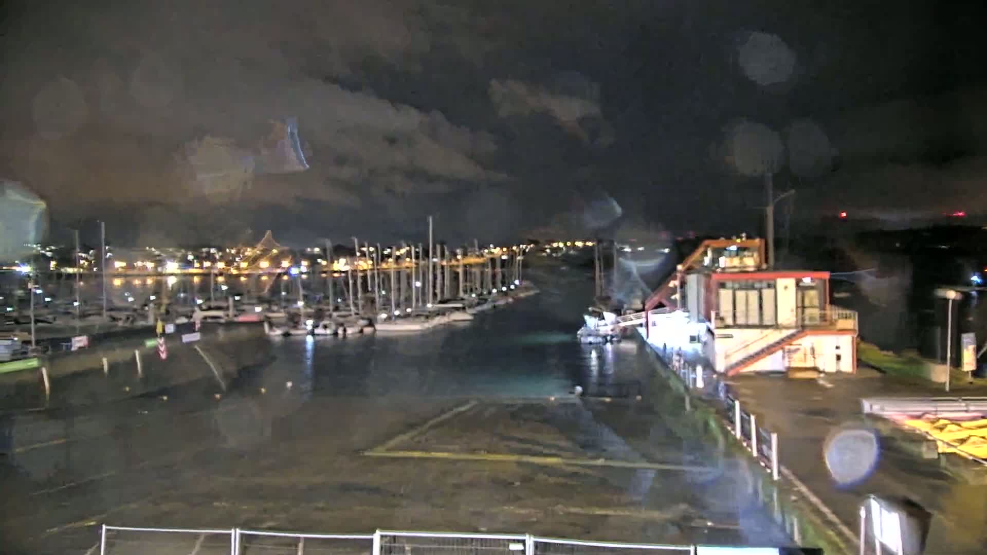 A nighttime view of a marina with many boats moored, under rainy conditions.