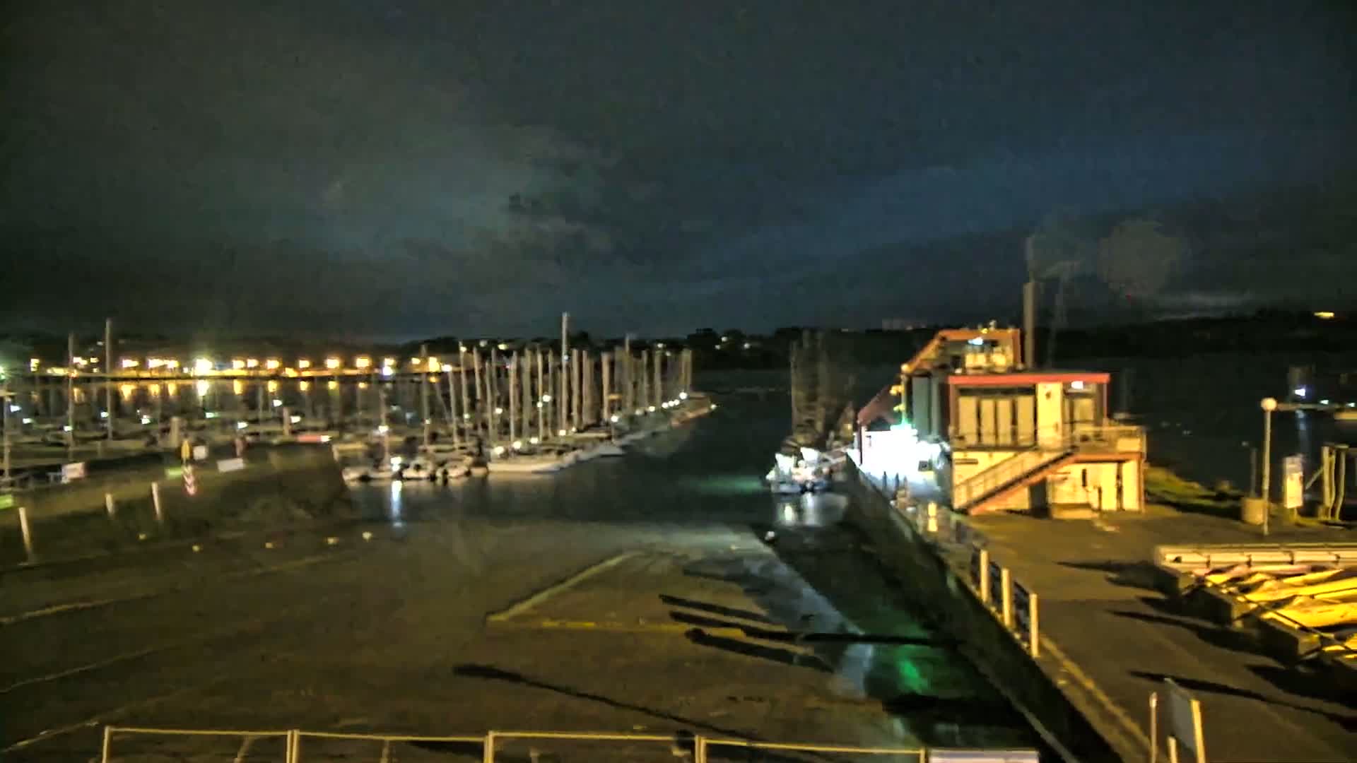 A nighttime view of a marina with numerous sailboats docked, a small building with lights on, and a dark, overcast sky.