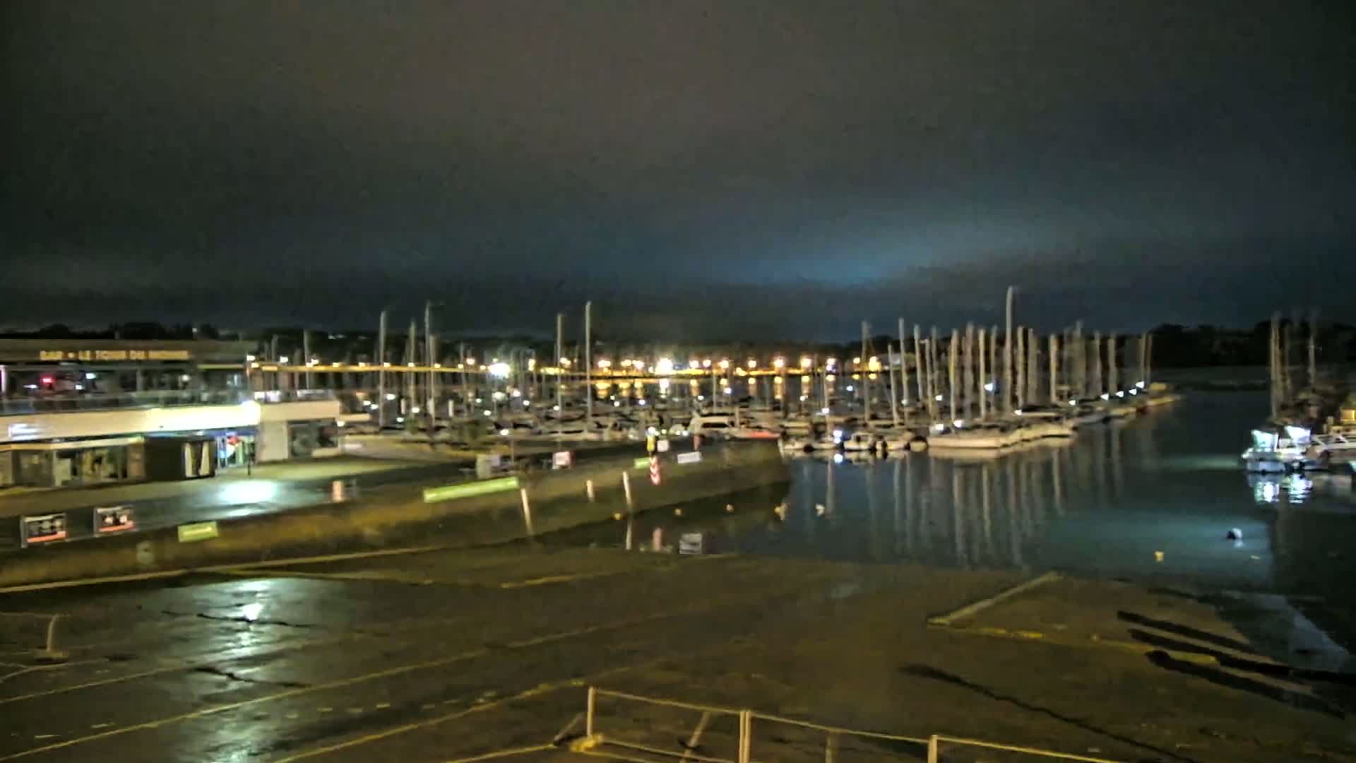 A marina at night under a dark, cloudy sky, filled with numerous sailboats and illuminated by lights along the docks and buildings.