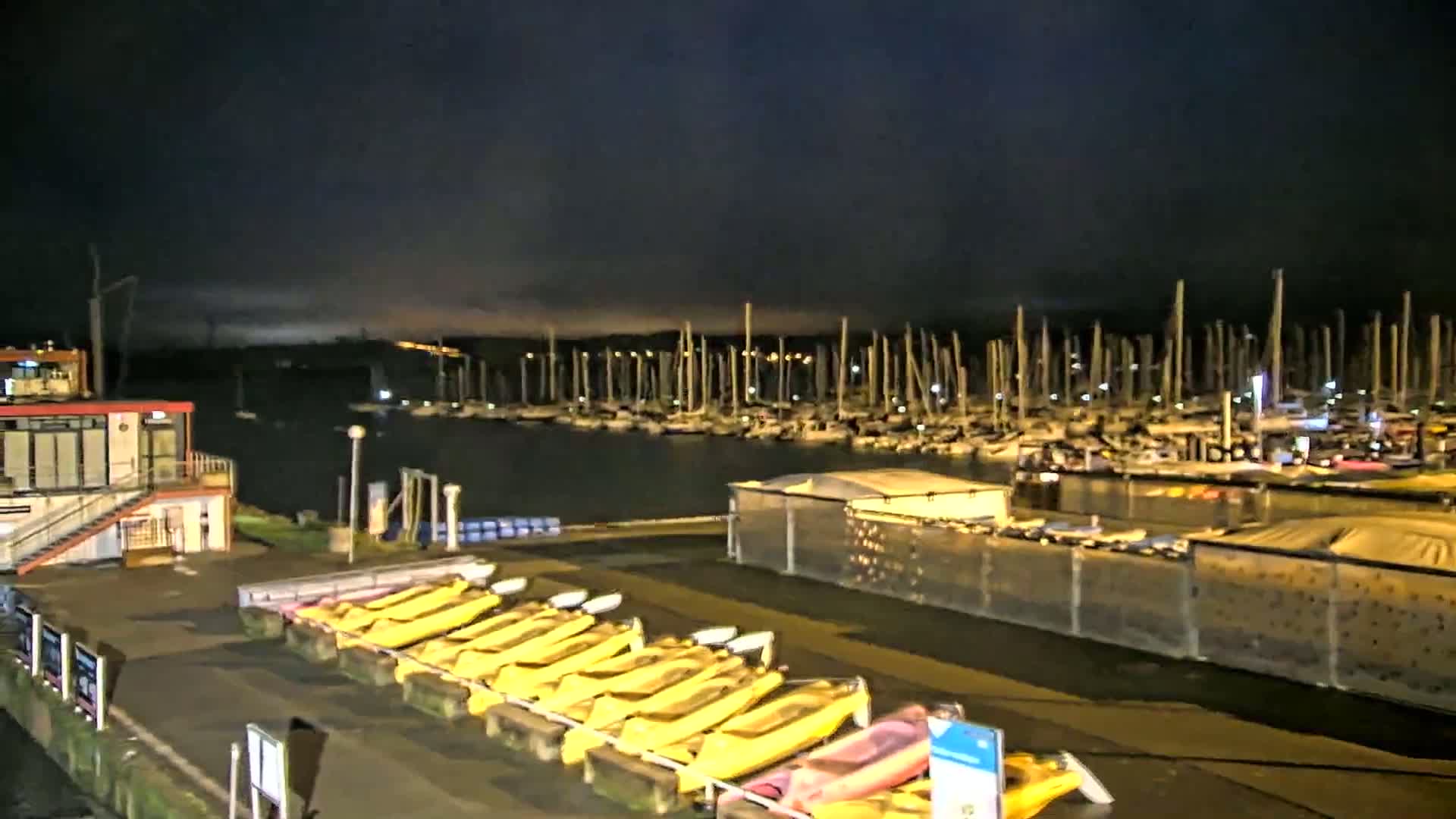 A nighttime view of a marina with numerous sailboats at dock, and a row of yellow kayaks near a small building, under a dark, cloudy sky.