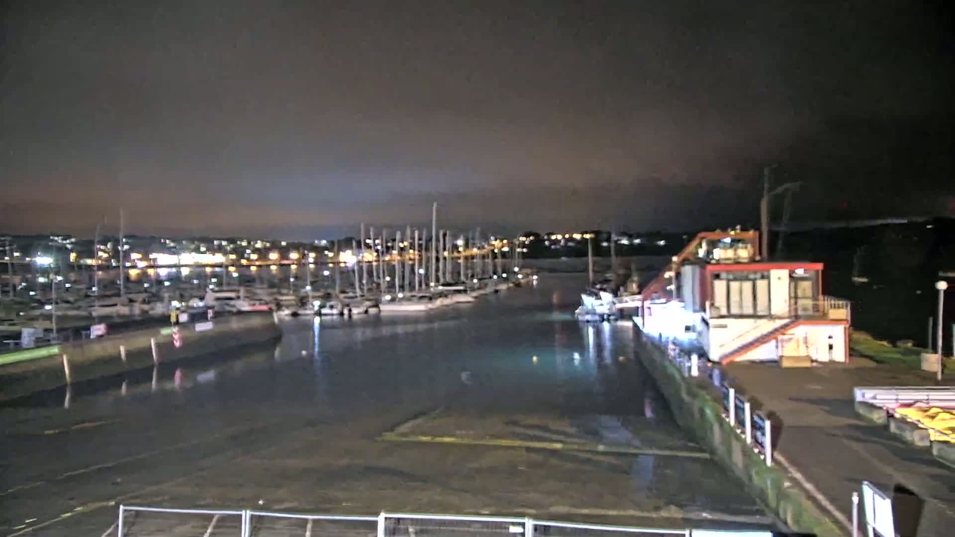 A nighttime view of a marina filled with docked sailboats, a small building, and a calm body of water under a dark sky.