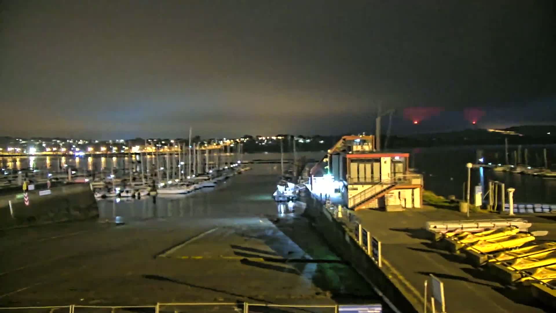 A marina at night, under a dark, cloudy sky with distant red lights visible on the horizon, shows numerous boats moored in the water, and a building with yellow boats on the dock.