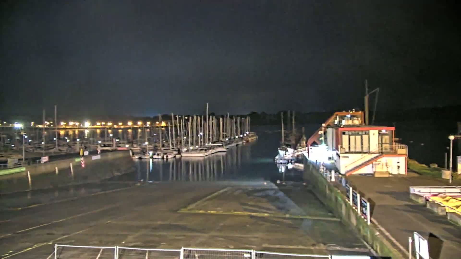 A marina at night with many sailboats docked, a small building with lights on, and a dark, calm body of water reflecting the lights.