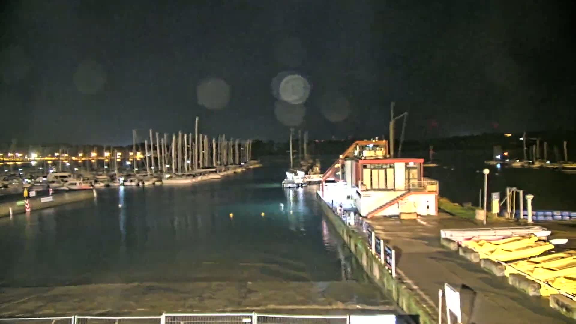 A nighttime view of a marina with numerous boats docked, a large barge or floating structure at a dock, and some rain or mist in the air.