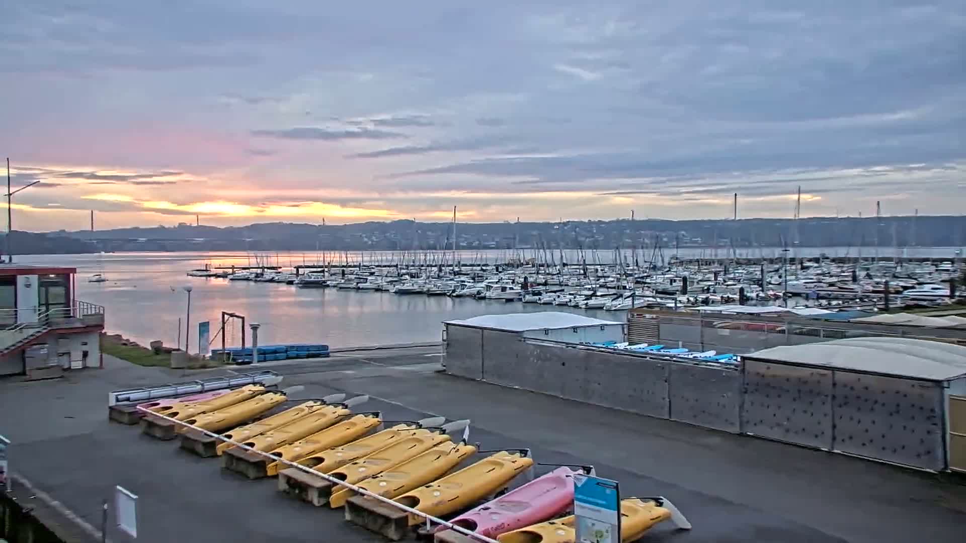 A calm harbor filled with numerous boats at sunset, with kayaks lined up on the shore.