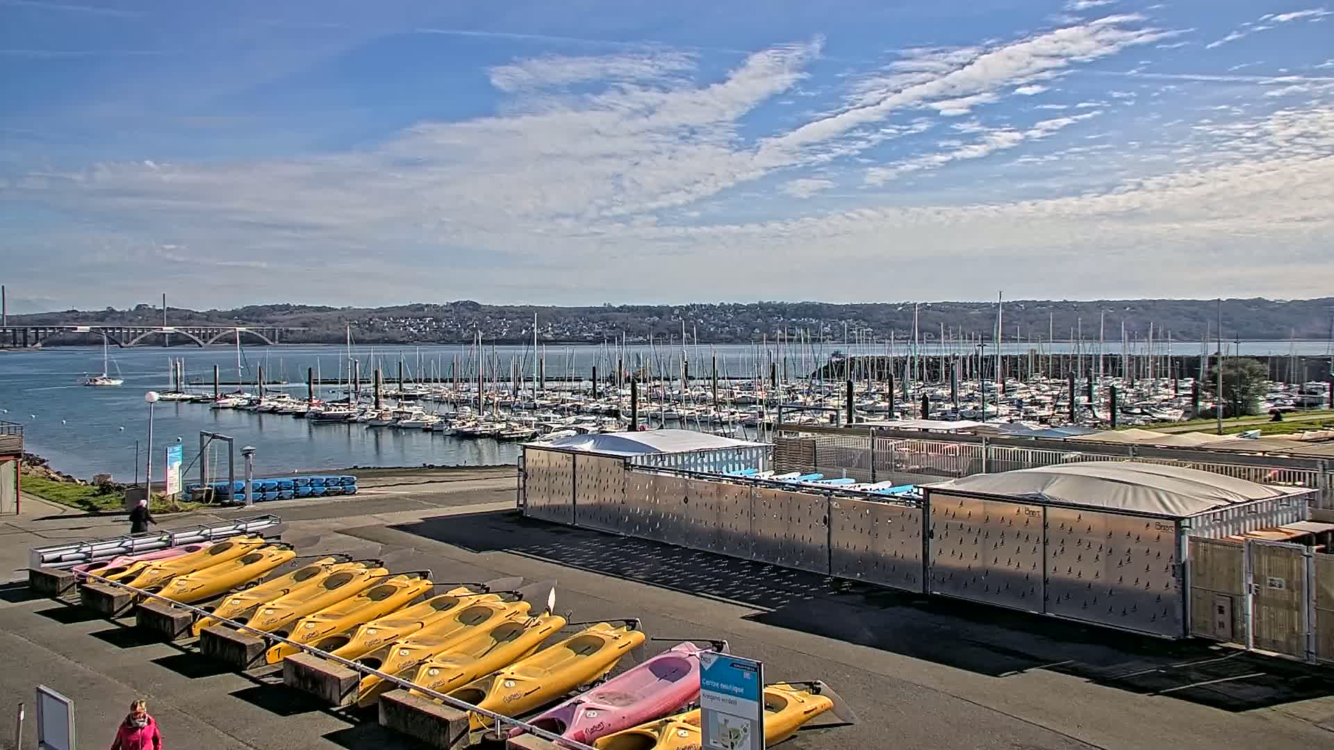 A sunny day overlooking a marina filled with sailboats, a bridge in the distance, and a row of kayaks parked on the shore.
