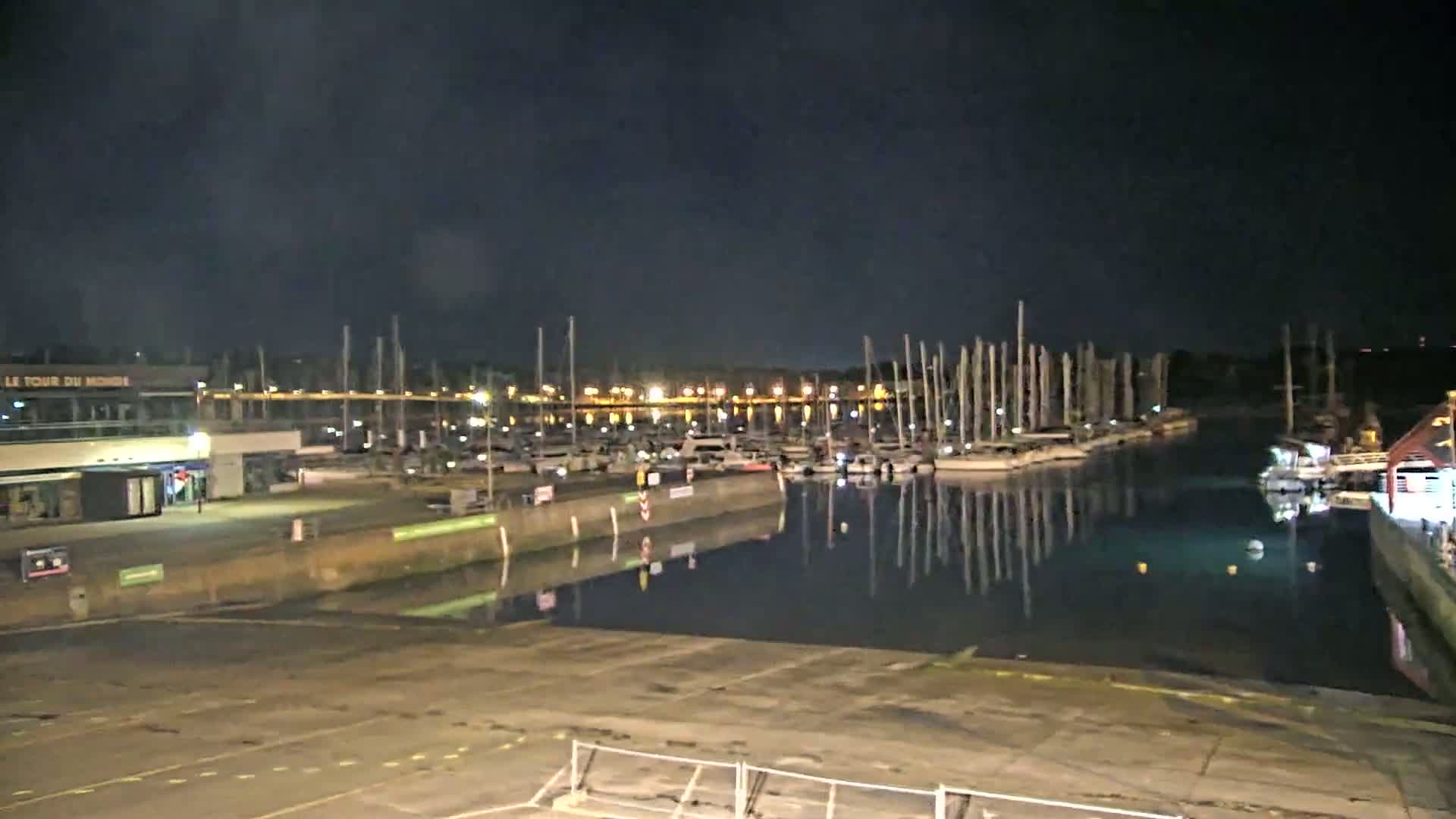 A nighttime view of a marina with numerous boats docked, their masts reflecting in the calm water under a dark sky.