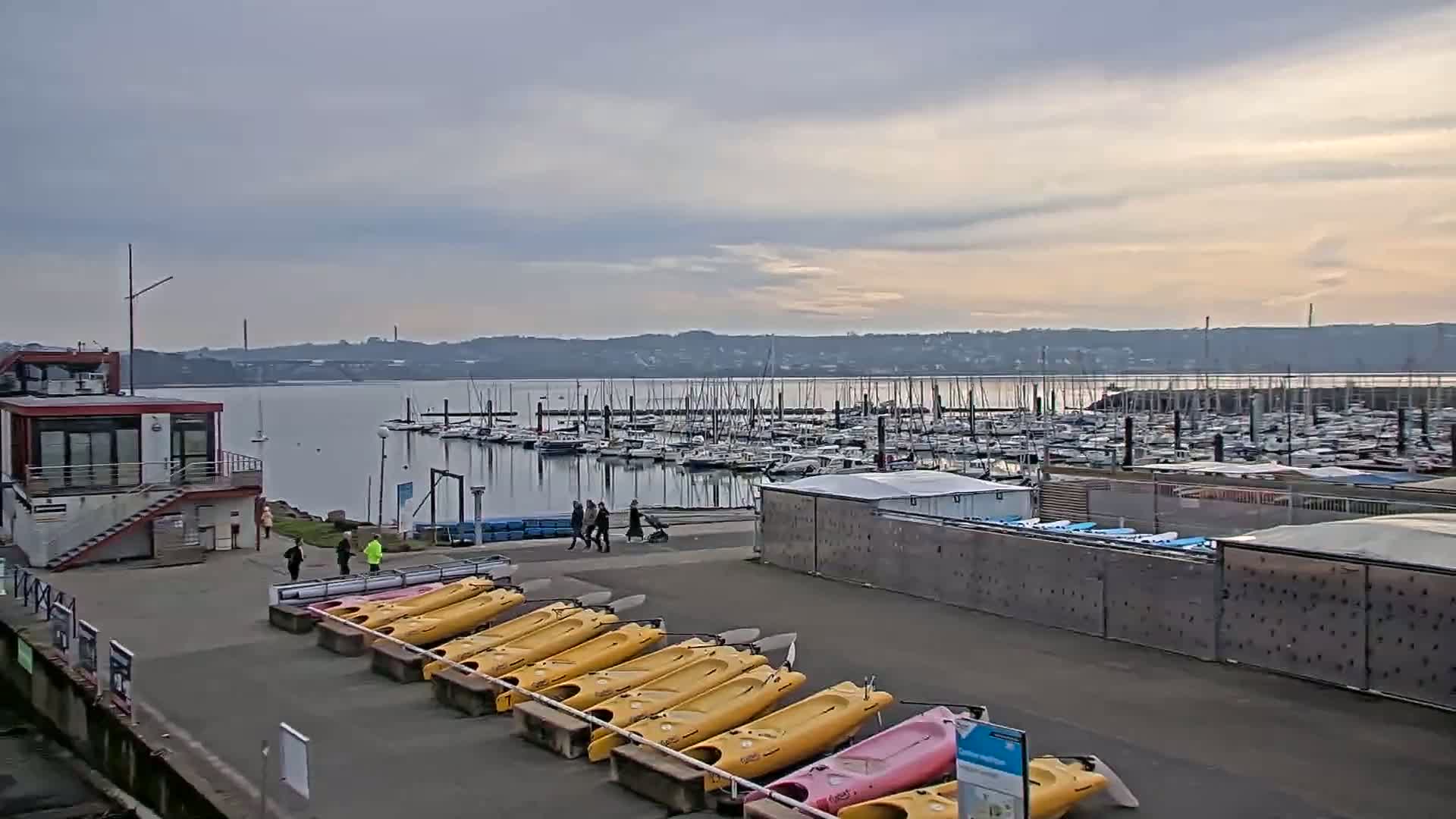 A marina with numerous sailboats and yachts in calm water under a cloudy sky,  kayaks are lined up on the shore near a small building.