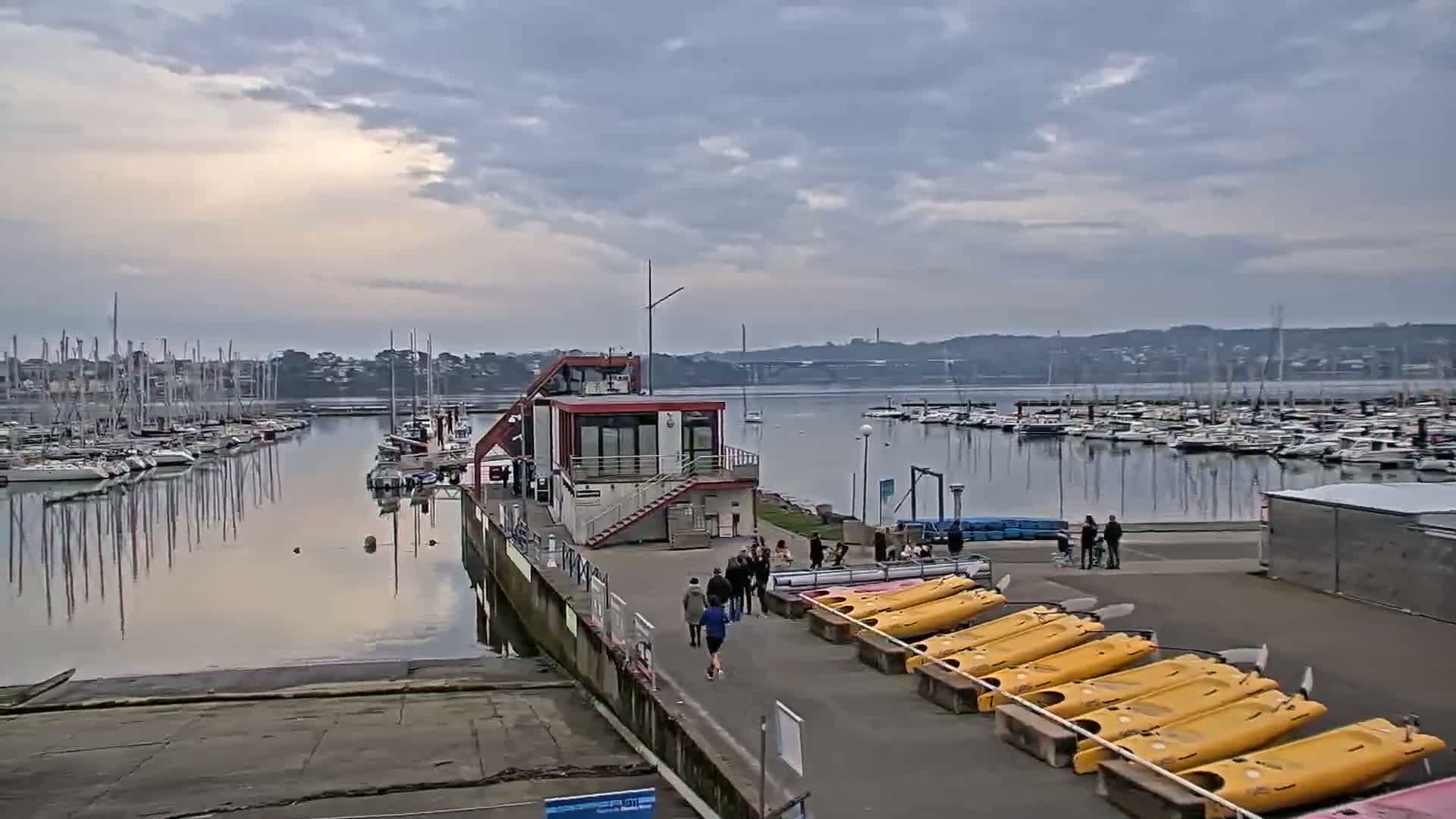 A marina filled with sailboats under a cloudy sky, with a small building and several people walking near rows of yellow kayaks.