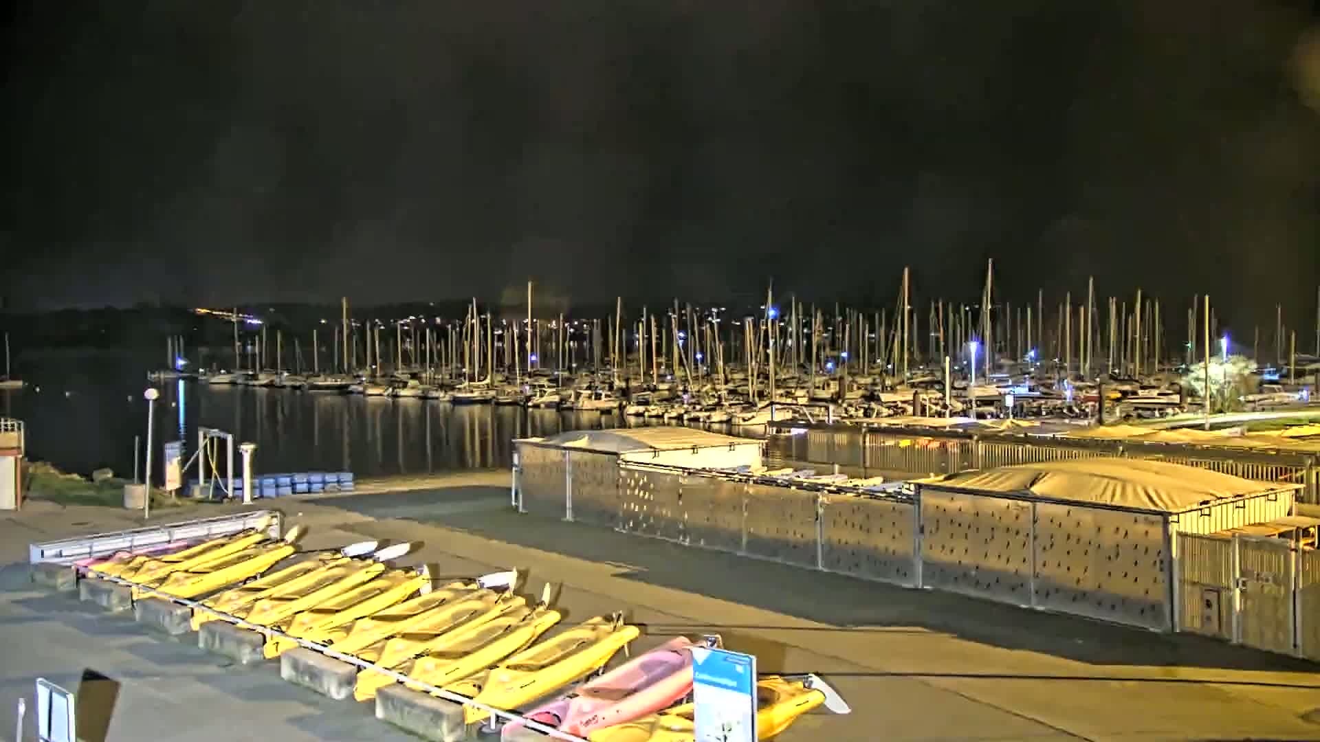 A nighttime view of a marina with numerous sailboats docked, and a row of kayaks stored on the shore under a dark sky.