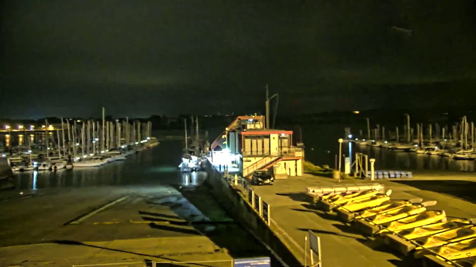 At night, a marina with numerous sailboats is illuminated, and several yellow small boats are lined up on a dock near a building.