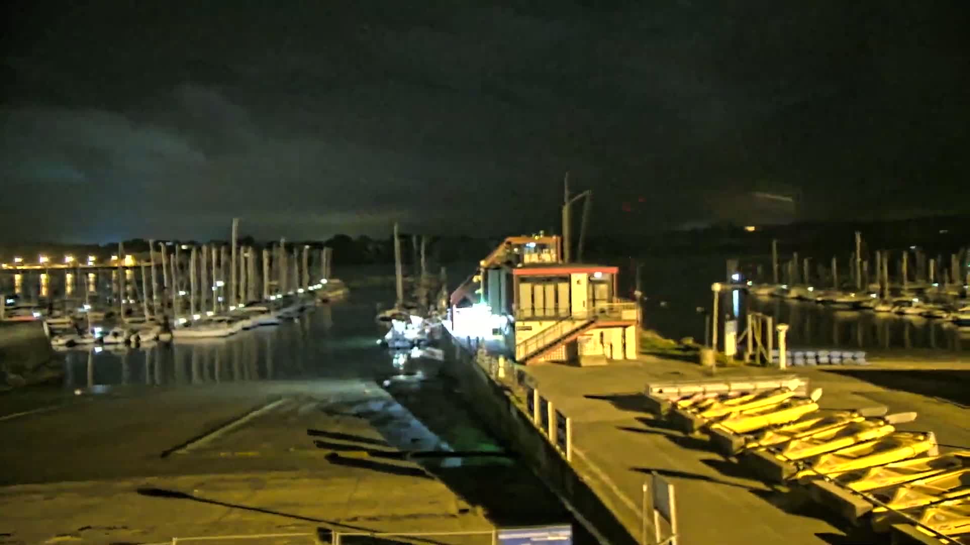 A nighttime view of a marina with numerous boats docked, a building with lights on, and several yellow kayaks lined up on a dock under a cloudy sky.