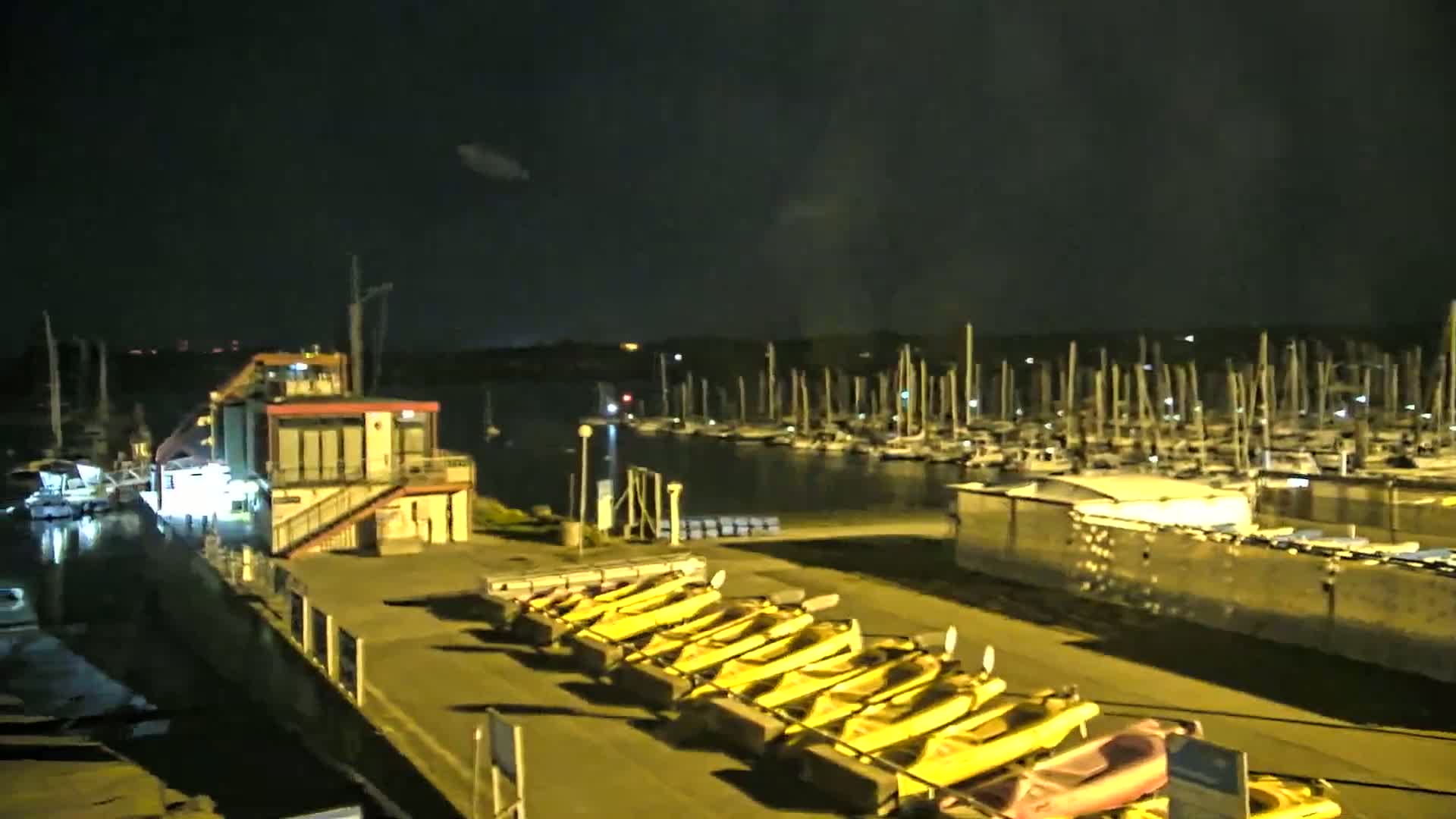 A marina at night, under a dark sky, shows numerous boats docked, a building with lights on, and a row of yellow kayaks lined up on the shore.