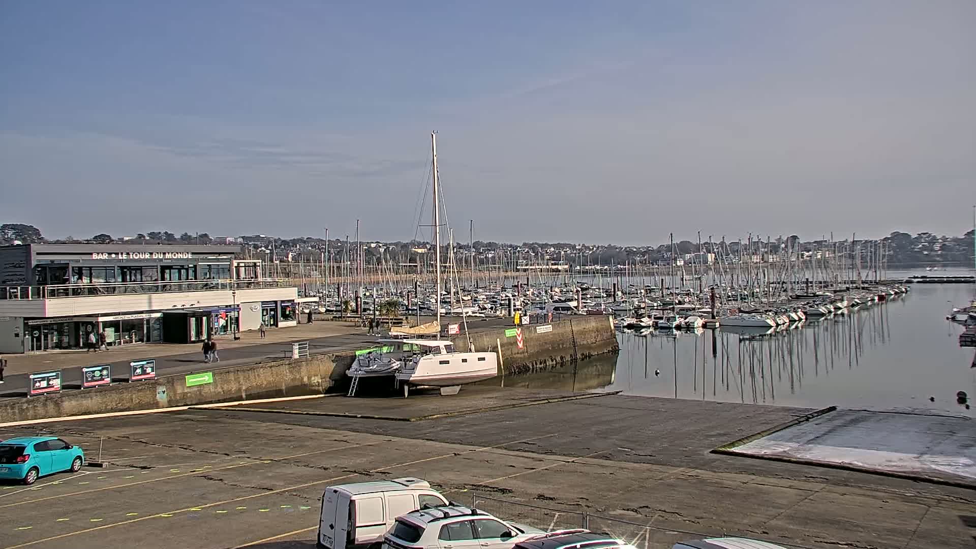 Under a clear blue sky, a marina filled with numerous sailboats is seen from a parking lot, with a building and several cars in the foreground.