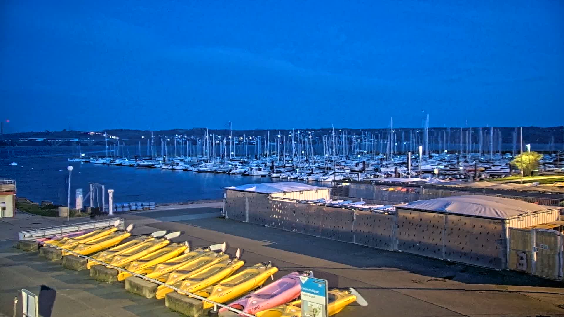 Under a twilight blue sky, numerous sailboats are docked in a marina, with a row of yellow kayaks resting on a shore nearby.