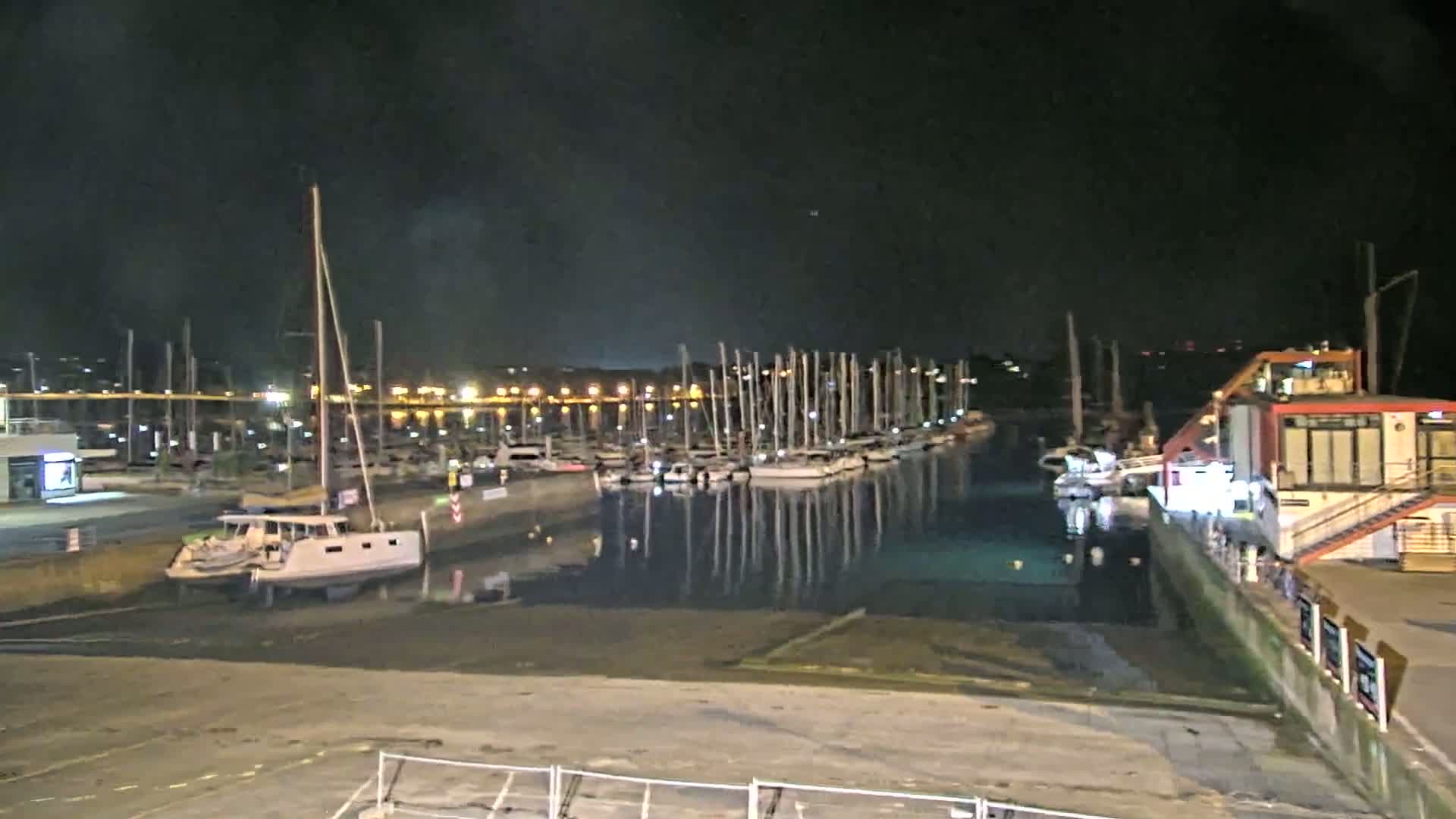 A nighttime view of a marina with numerous sailboats moored, a catamaran resting in shallow water, and a building with lights on.