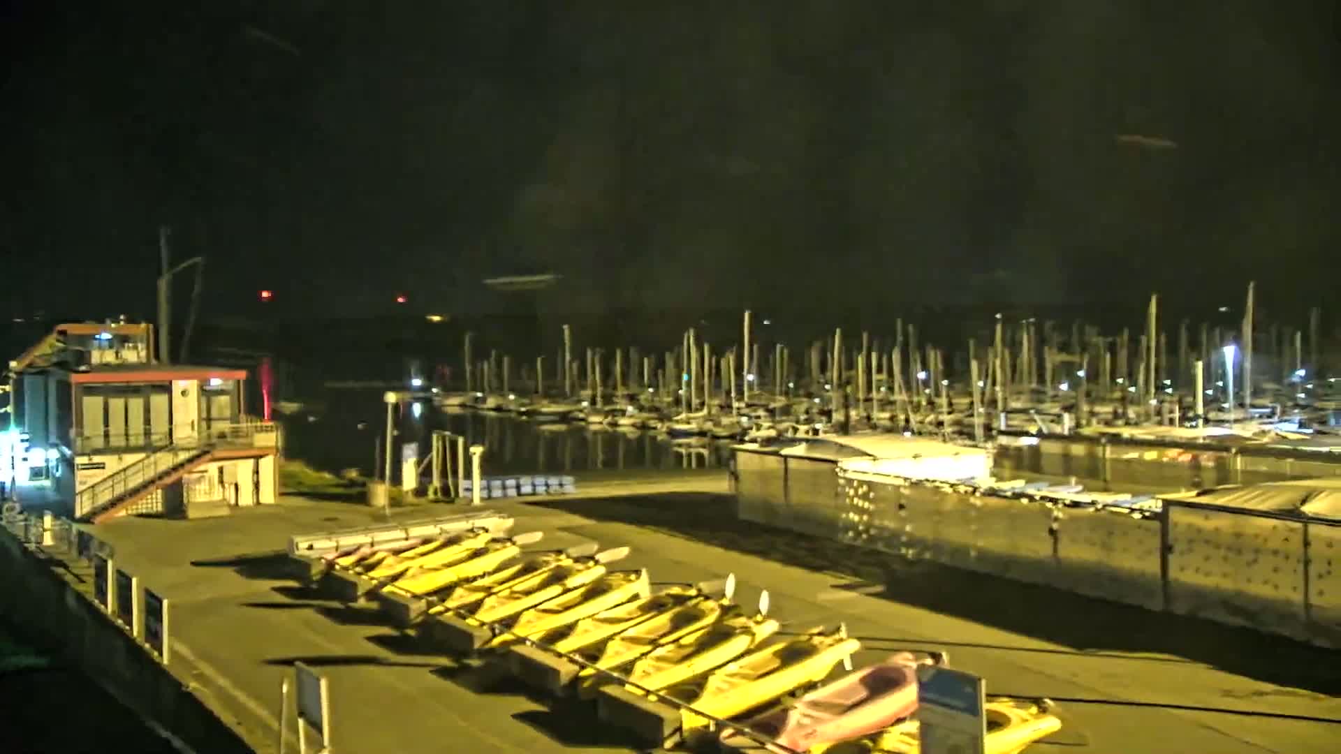A nighttime view of a marina with numerous docked sailboats and a row of yellow kayaks lined up on a dock near a small building.