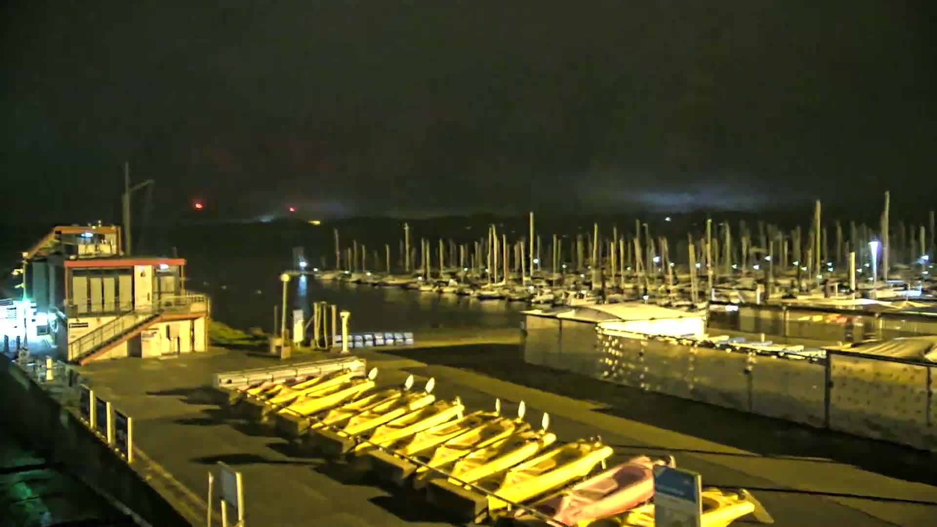 A marina at night with many docked sailboats is seen under an overcast sky, with a row of yellow small boats parked nearby a small building.