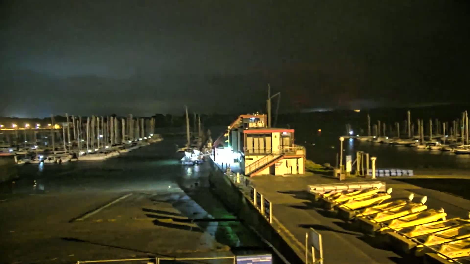 A marina at night with numerous sailboats docked, a building with lights on, and several yellow boats lined up on a dock under a dark, cloudy sky.
