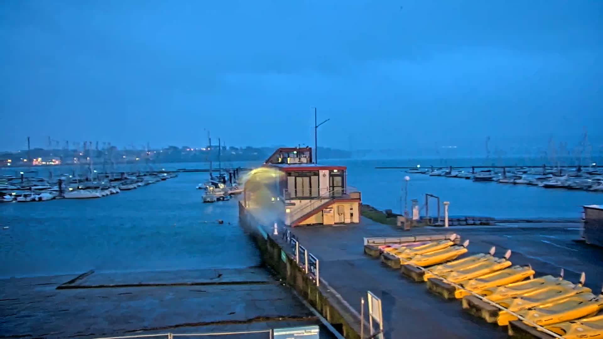 A marina at dusk with choppy water splashing against a building on a pier, and numerous boats docked nearby.