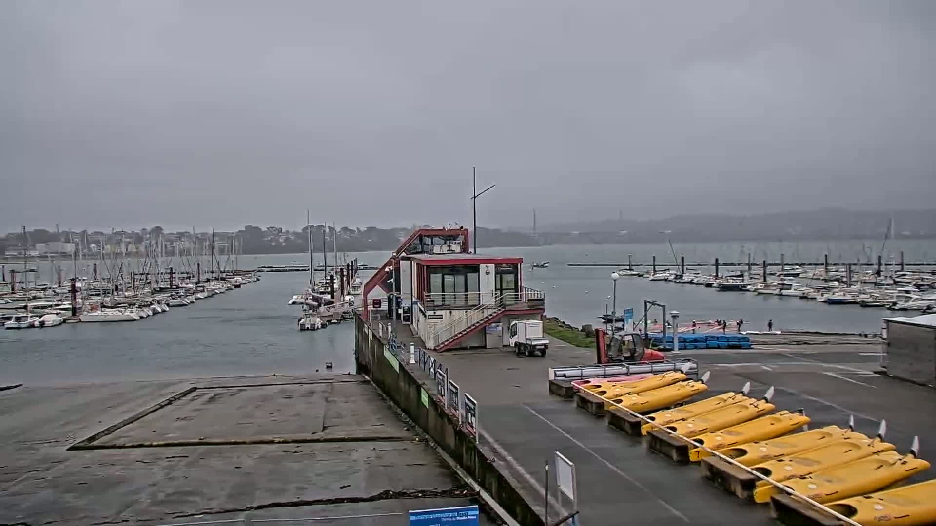 A marina filled with sailboats on a cloudy day, featuring a small red building and numerous yellow kayaks lined up on the shore.