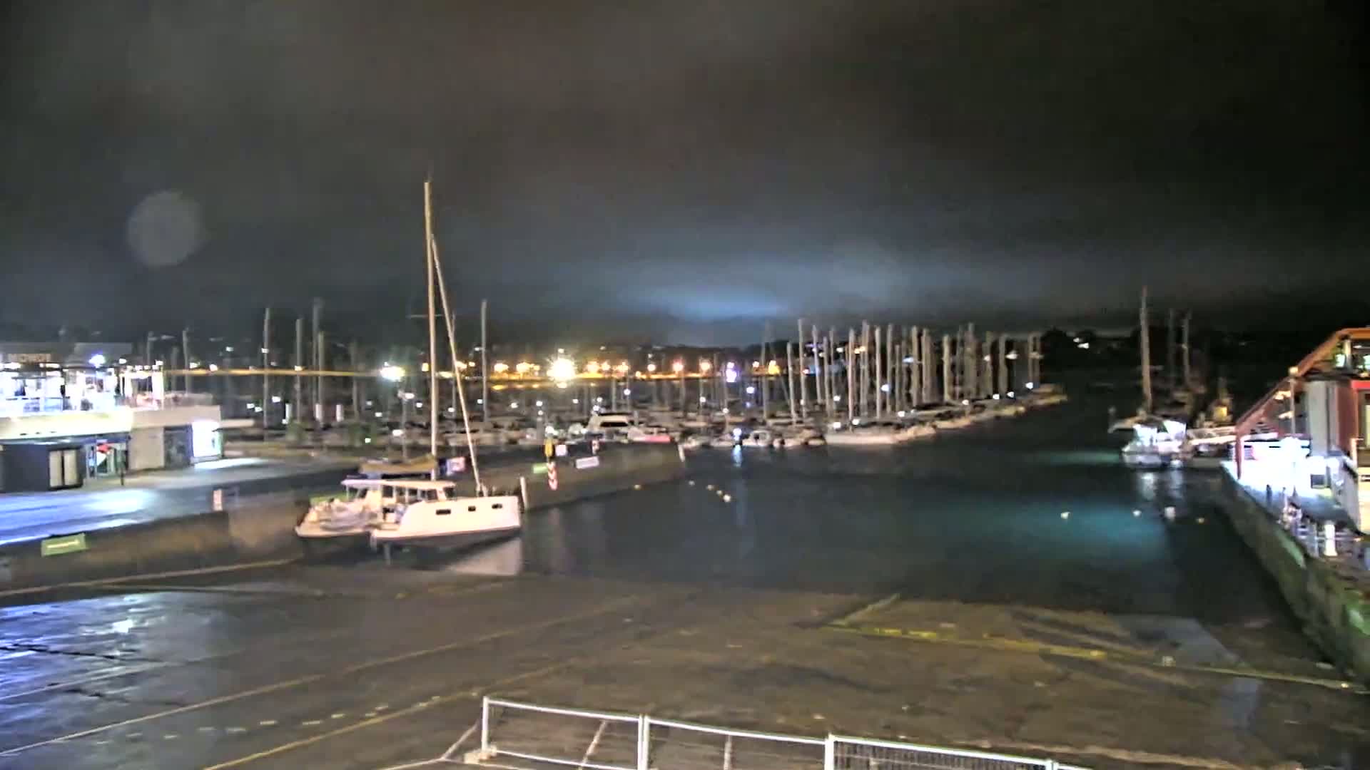 A marina at night under a dark, cloudy sky, filled with numerous sailboats and motorboats docked along the waterfront.