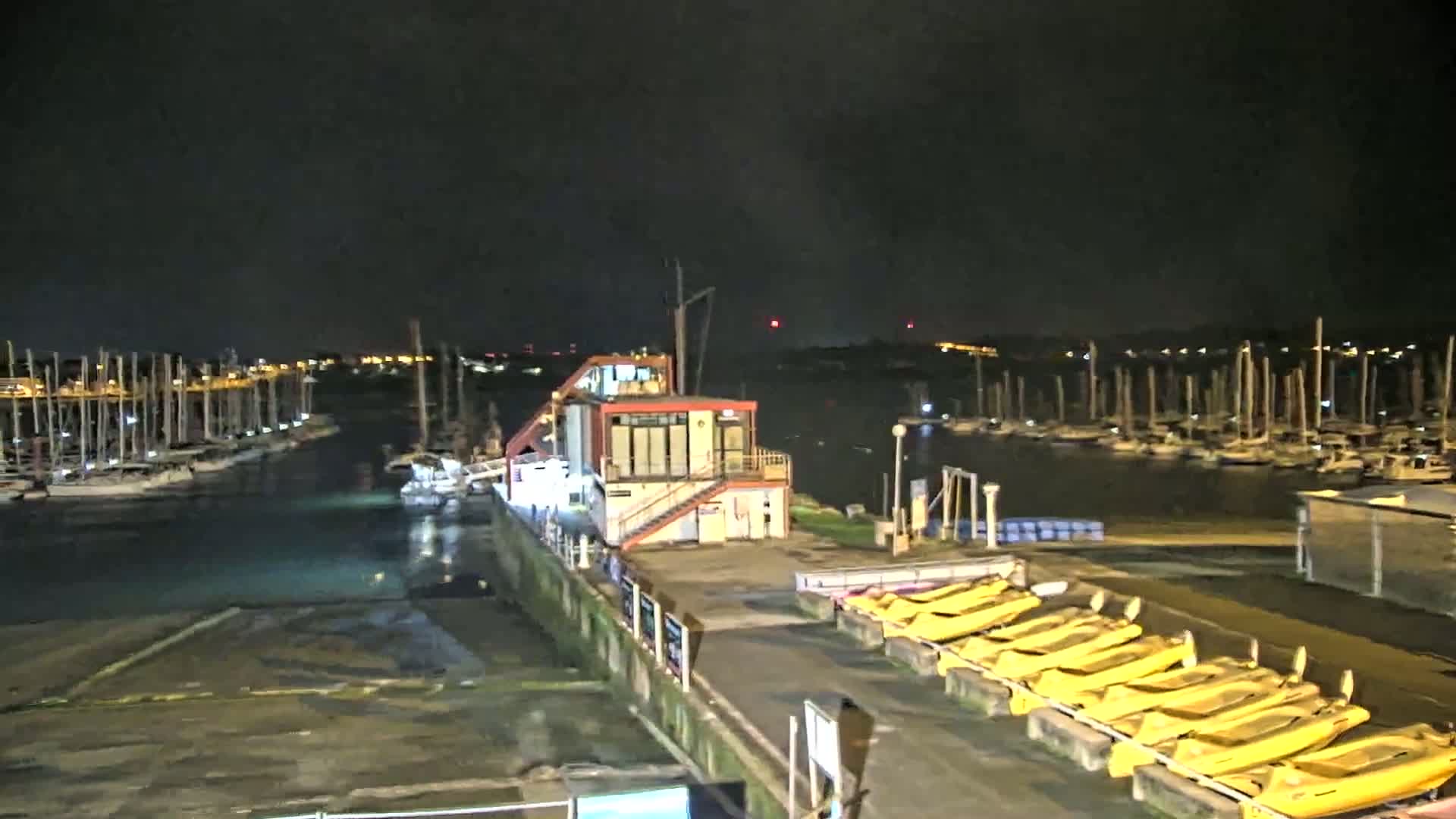 At night, a marina filled with sailboats is shown, with a small building and a line of yellow boats docked nearby.