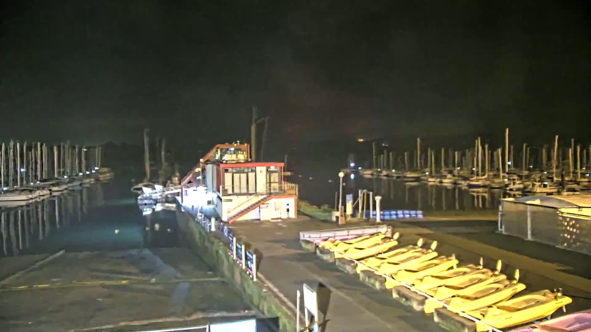 At night, a marina is shown with many boats docked, a small building, and several yellow boats lined up on the shore.
