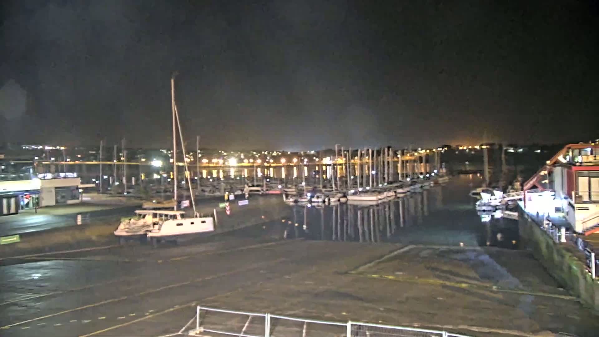 At night, a marina shows numerous boats docked along a calm waterway under a dark sky.