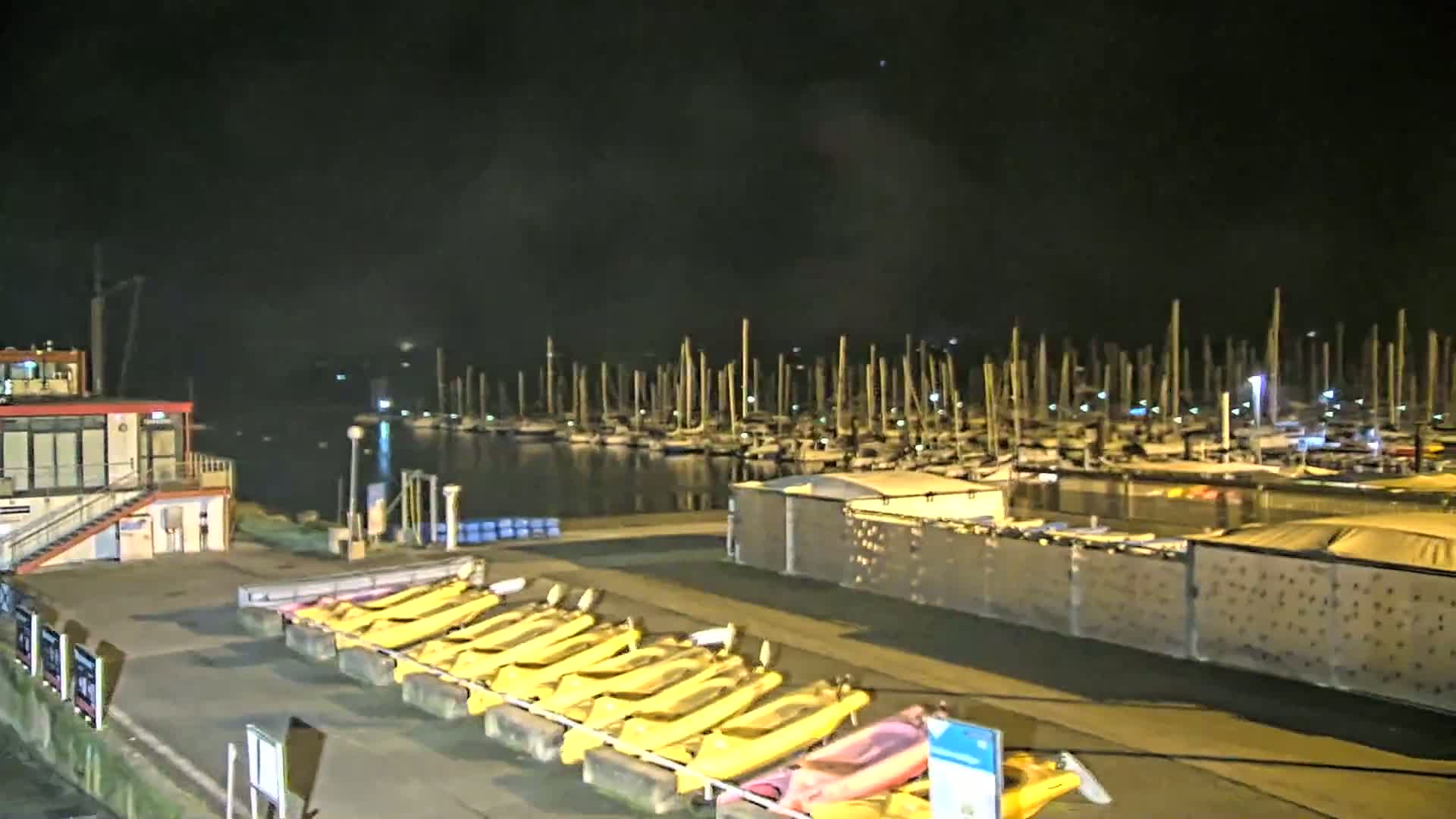 At night, a marina filled with docked sailboats is visible across a calm body of water from a vantage point showing a row of yellow kayaks and a small building.