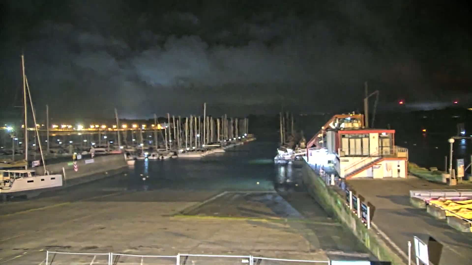 A marina at night under a cloudy sky, filled with numerous sailboats docked along a pier, next to a small building.