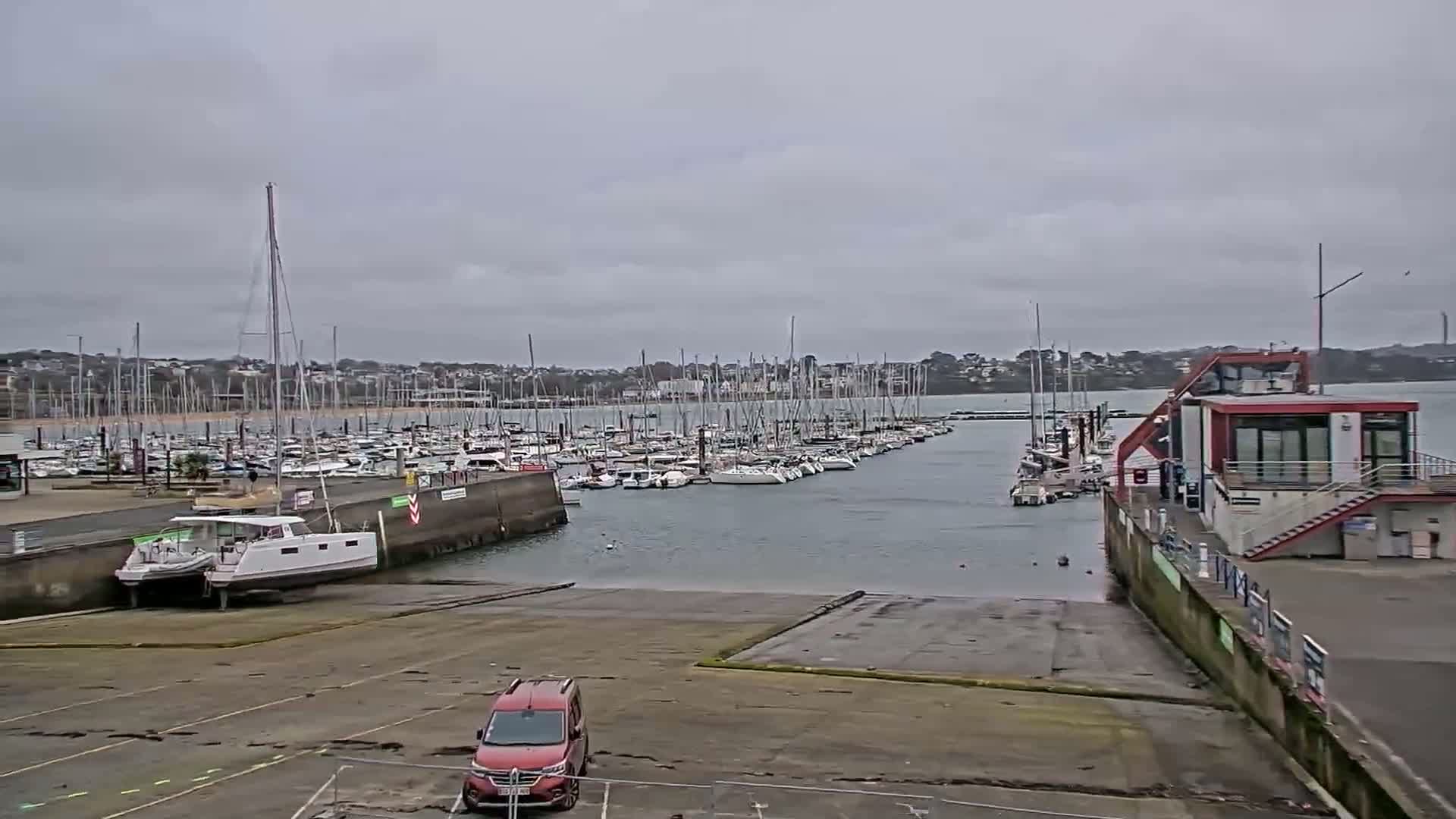 A marina filled with numerous sailboats is viewed from a boat ramp on an overcast day.