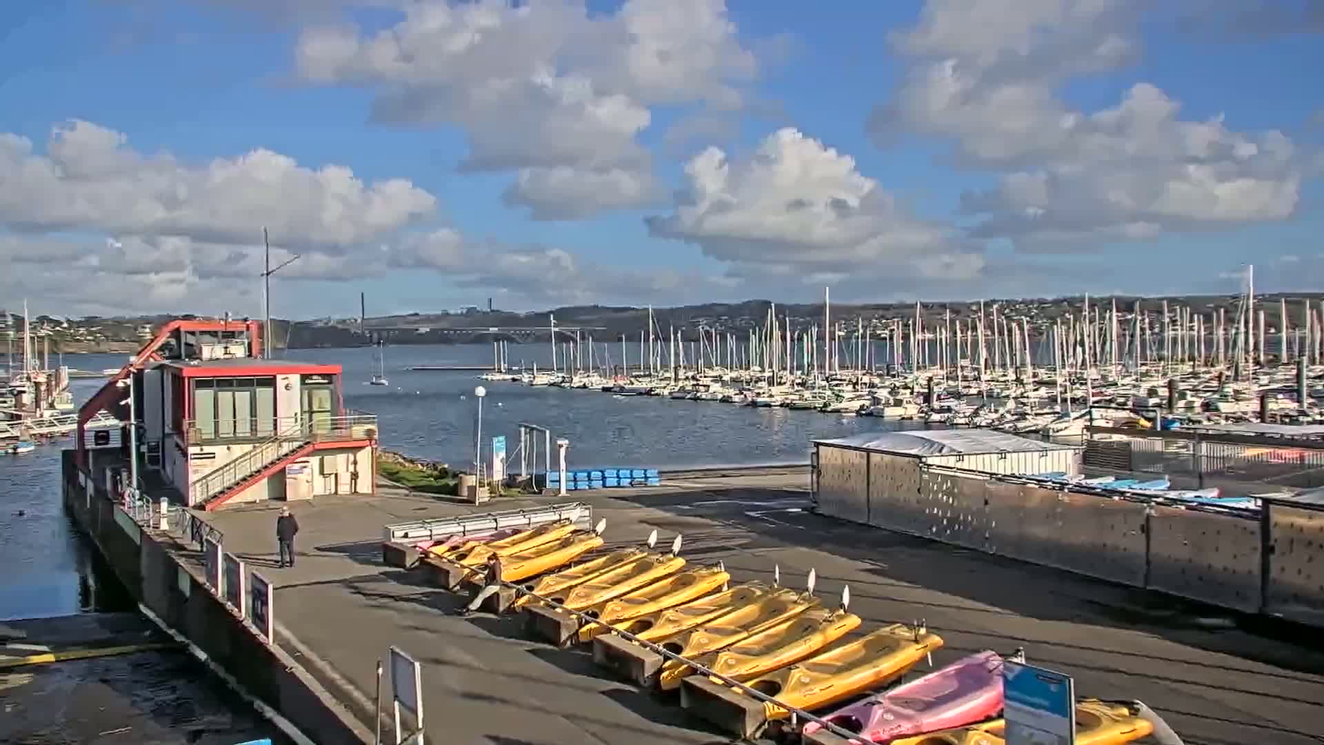A marina filled with sailboats under a partly cloudy sky, with kayaks lined up on a dock in the foreground and a small building nearby.