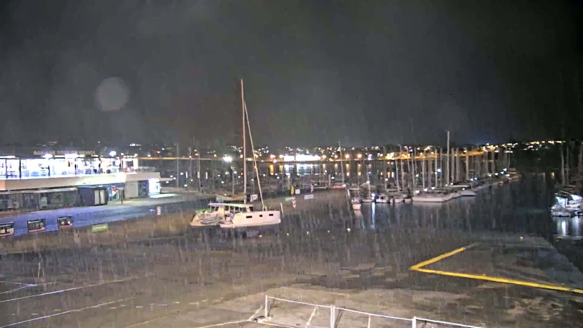 A nighttime marina scene shows rain falling on a mostly empty harbor, with a few boats moored and a catamaran near the shore, under a dark sky.