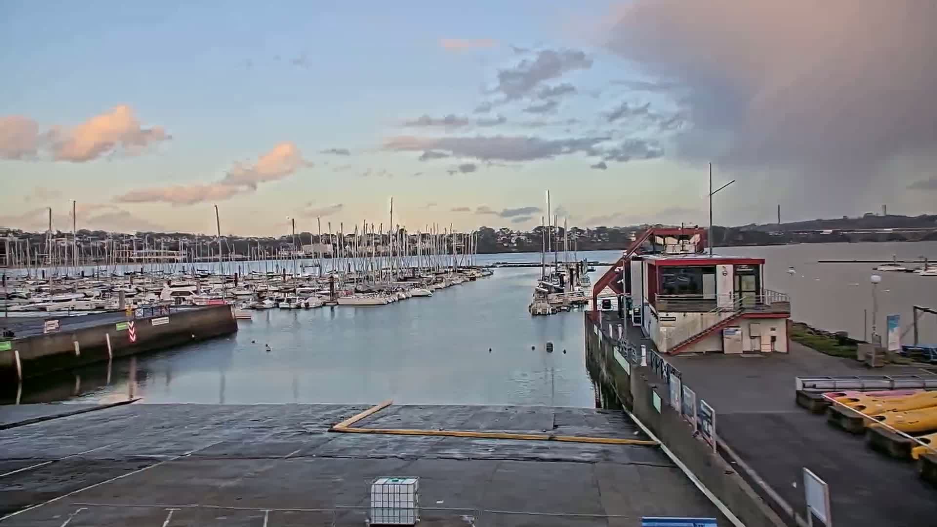 A marina filled with sailboats under a partly cloudy sky is seen from a dock with a small building and kayaks.