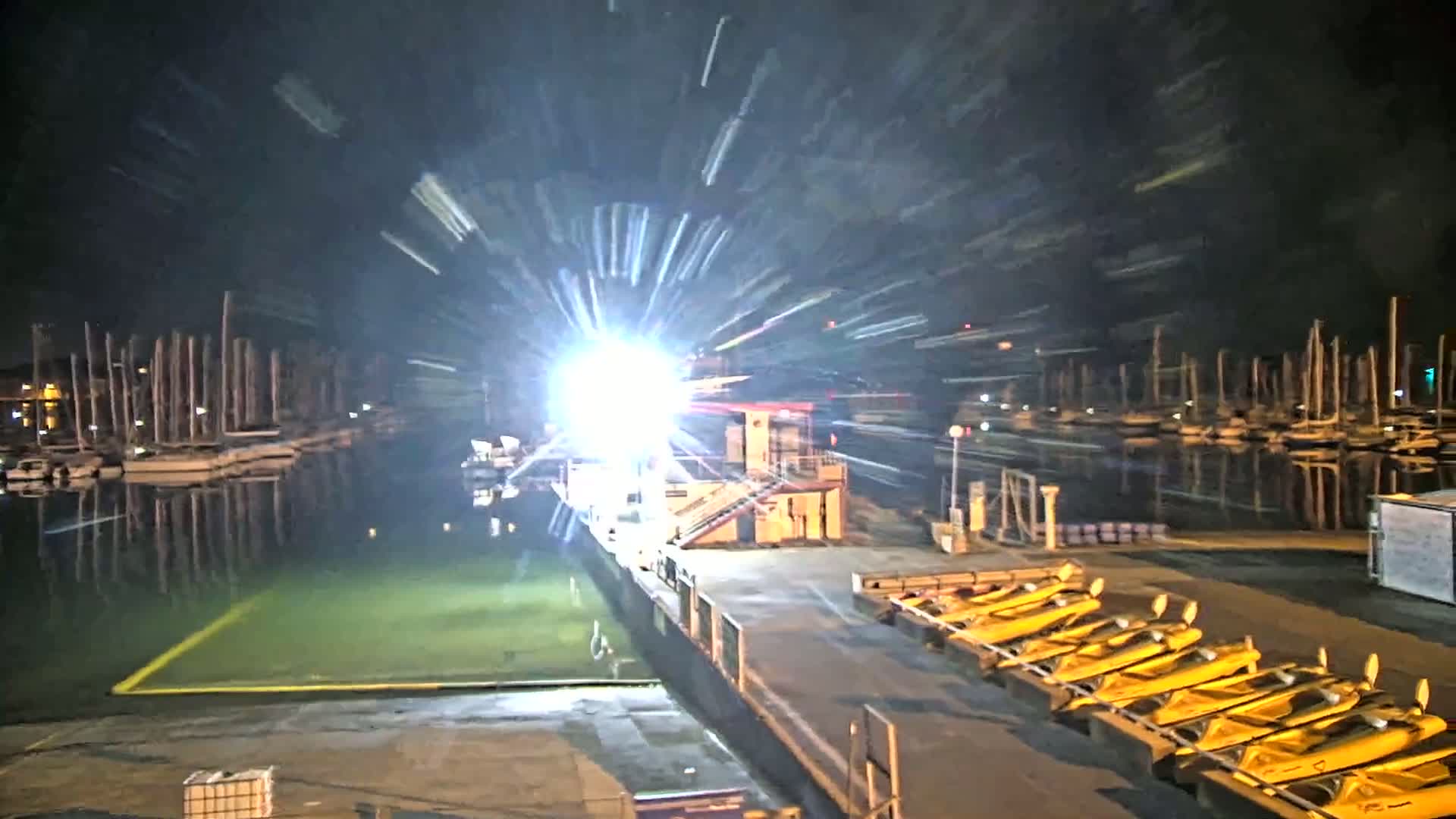 A brightly lit dock at night, with numerous boats moored in the water, and several yellow kayaks lined up on the dock.