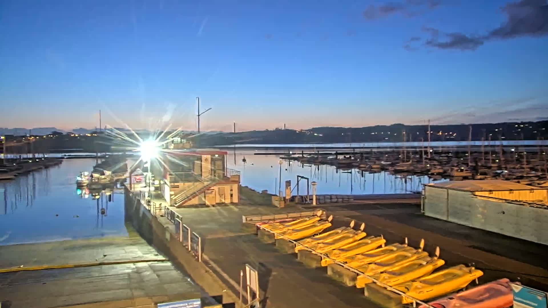 A marina at dawn shows numerous boats moored in calm water, with yellow kayaks lined up on the shore near a small building.