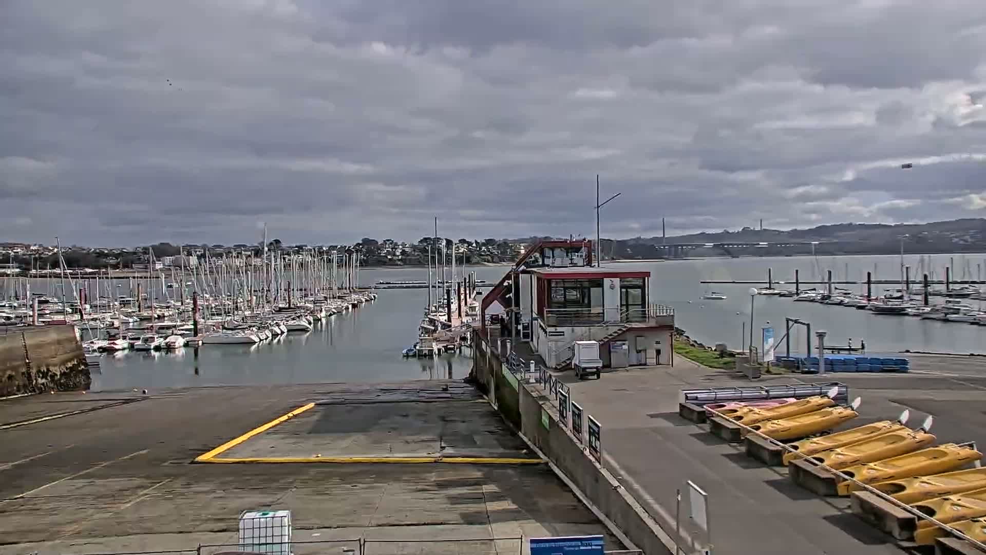 A marina filled with sailboats under a cloudy sky, with a small building and several kayaks visible near the launch ramp.