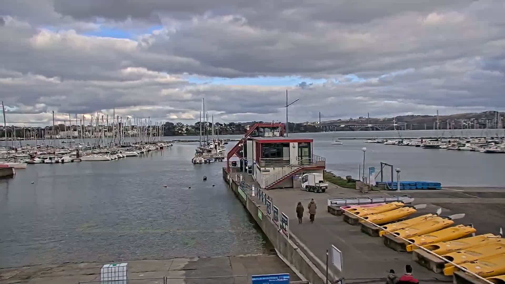 A marina filled with sailboats under a cloudy sky, with a small building and kayaks visible in the foreground.