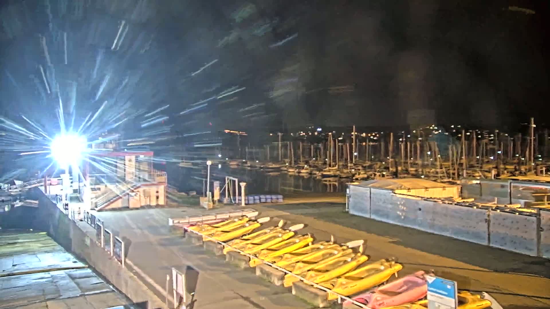 A nighttime view of a marina with numerous boats docked, a bright light shining, and a row of yellow kayaks lined up on the shore.