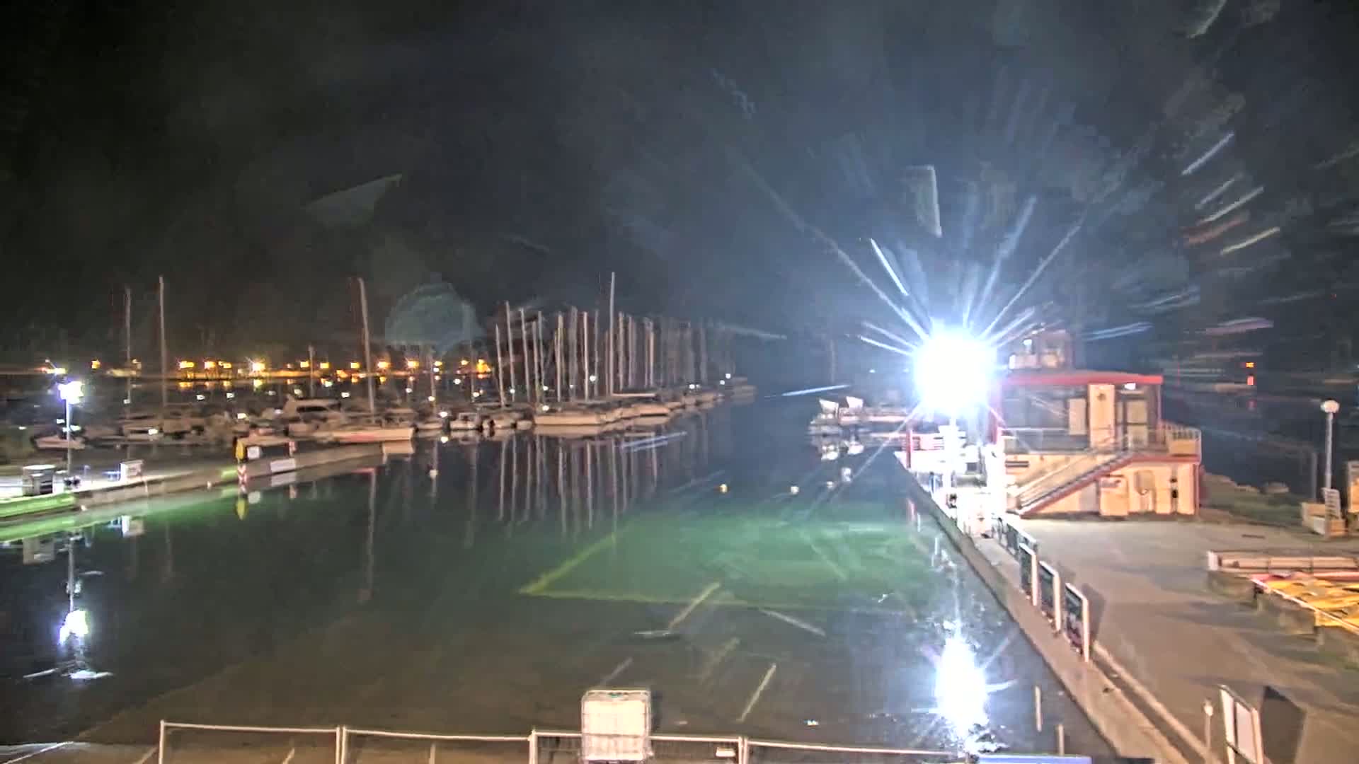 At night, a marina with numerous boats is brightly lit, its calm waters reflecting the lights and boats.