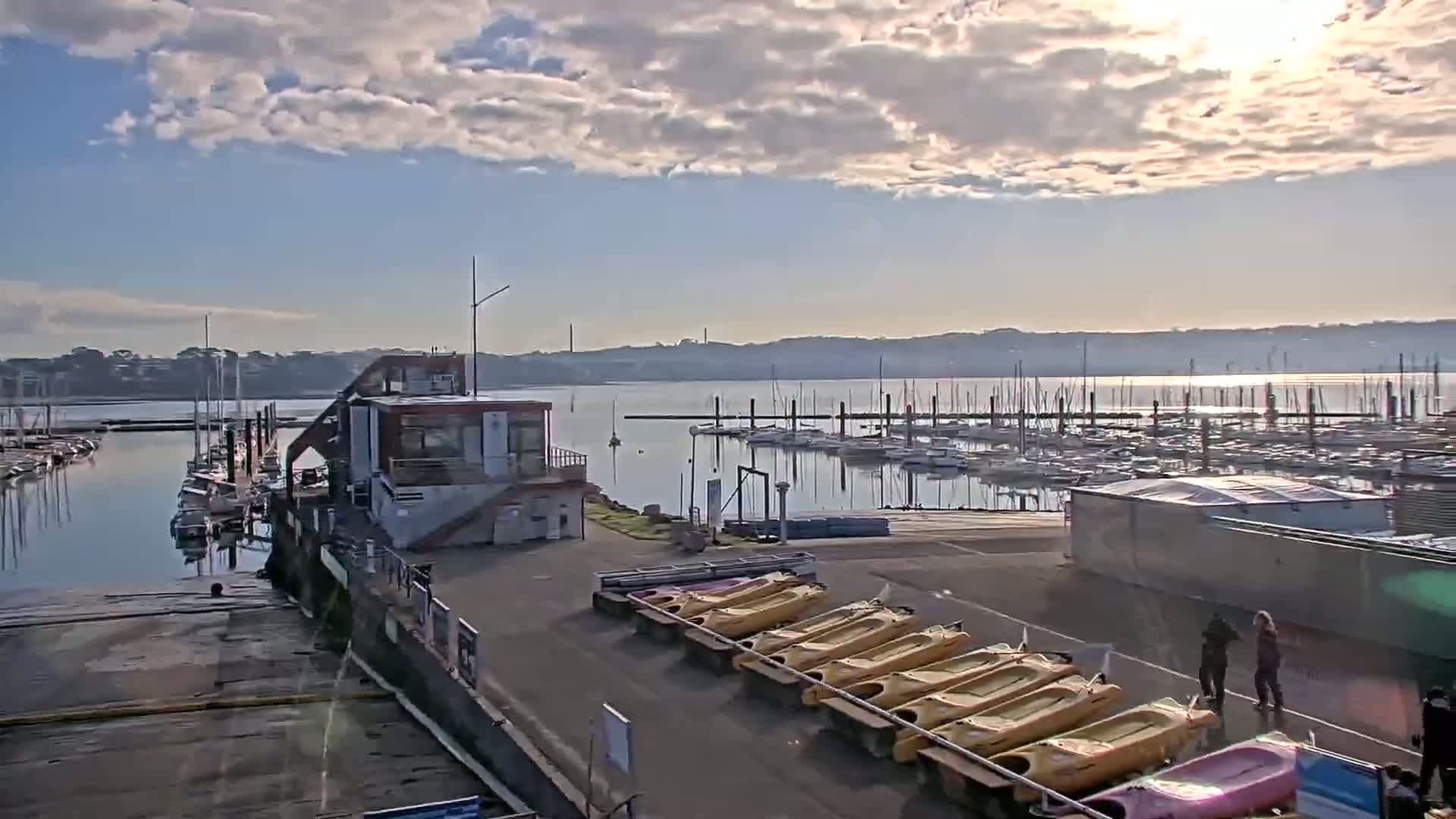 A marina with numerous sailboats docked, a small building, and several kayaks on trailers is visible under a partly cloudy sky.