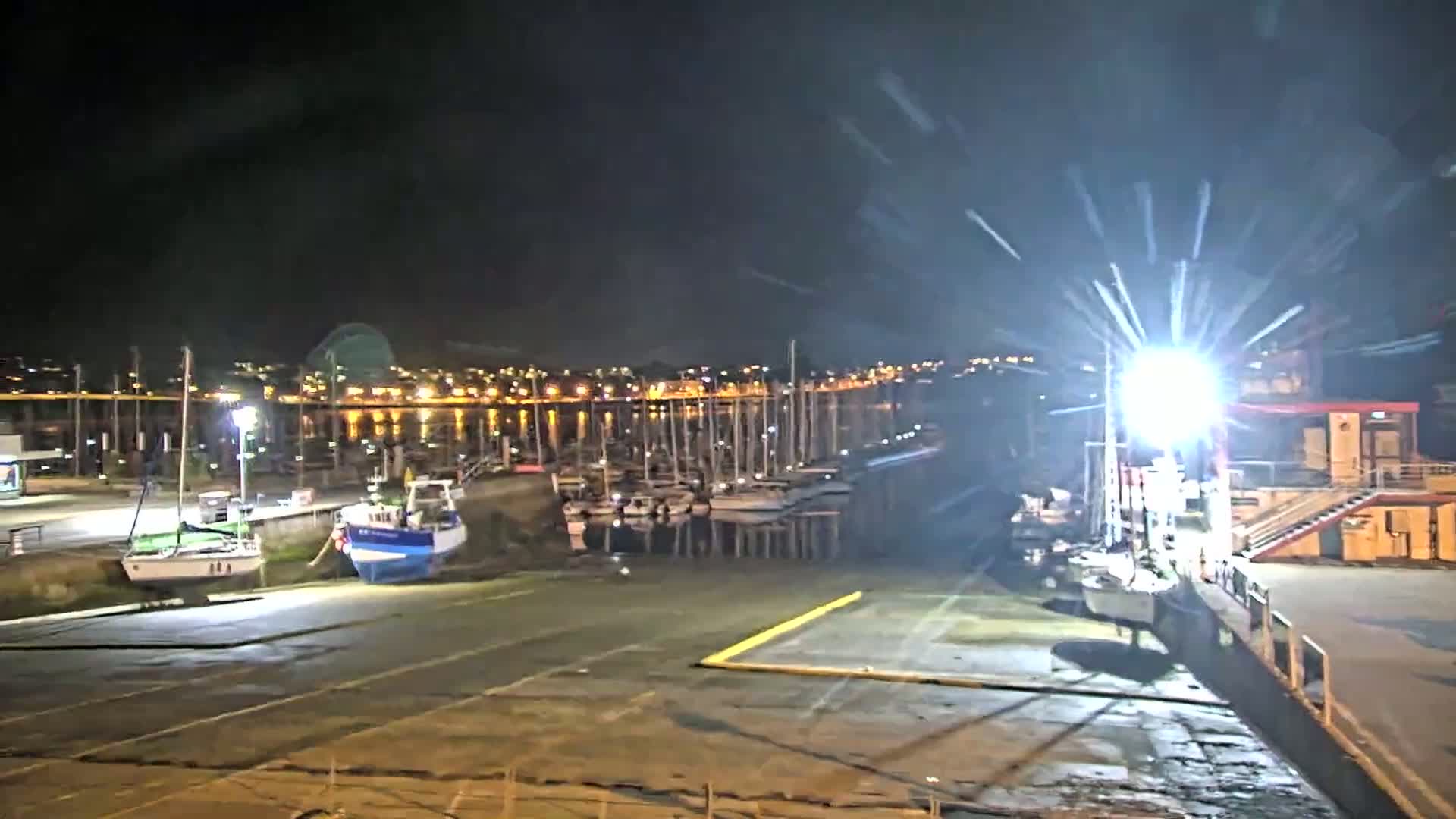 At night, a harbor is illuminated, showing several boats docked along a pier, with a brightly lit area near a building and a city skyline visible in the background.