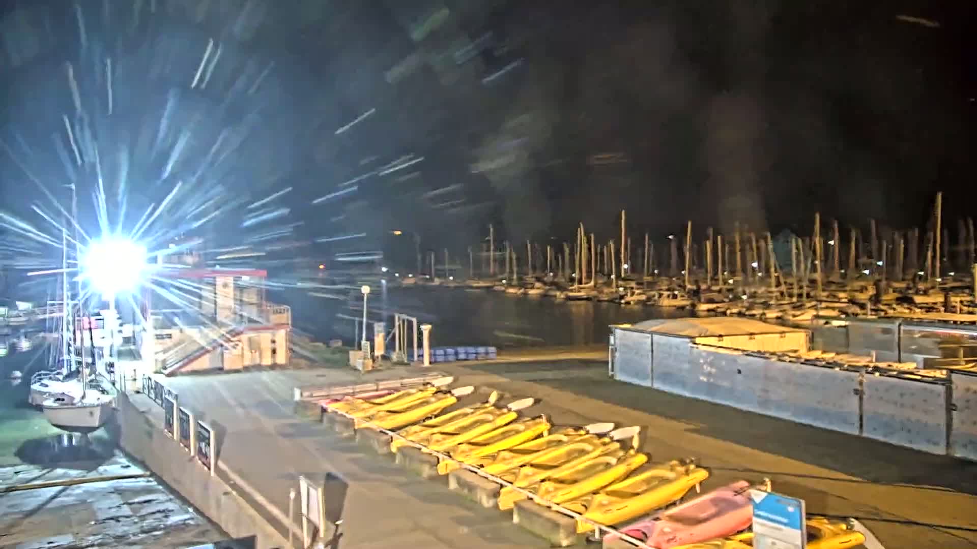 A nighttime view of a marina with numerous sailboats docked, a bright light shining, and a row of yellow kayaks lined up on the shore under a slightly blurry sky.