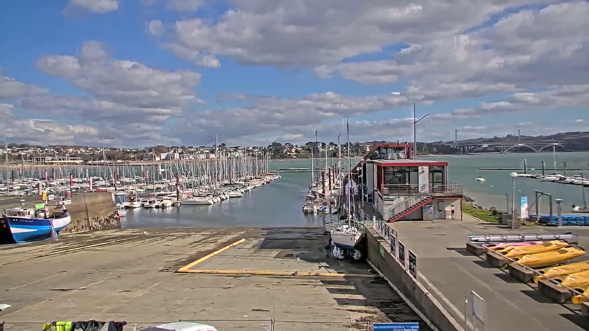 A marina filled with sailboats under a partly cloudy sky, with a waterfront building and kayaks nearby.