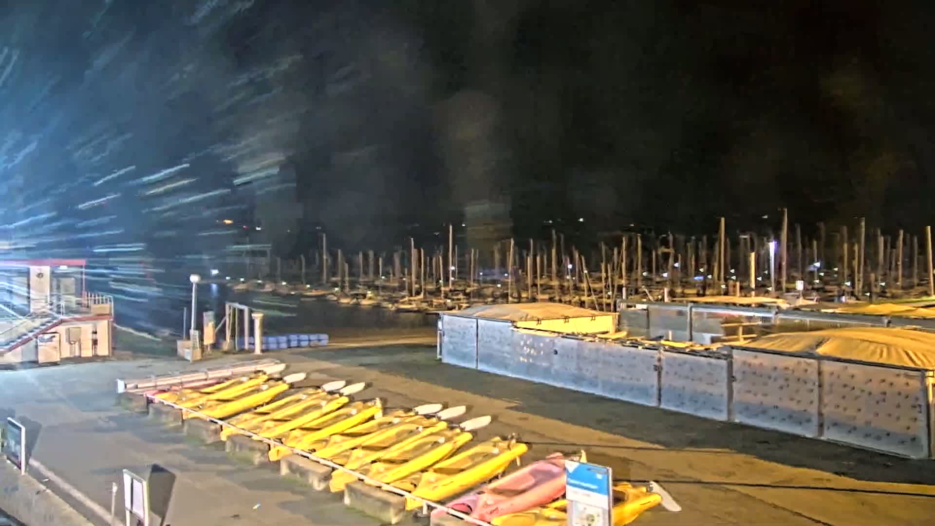At night, under a dark, windy sky, numerous yellow kayaks are lined up near a marina filled with sailboats.