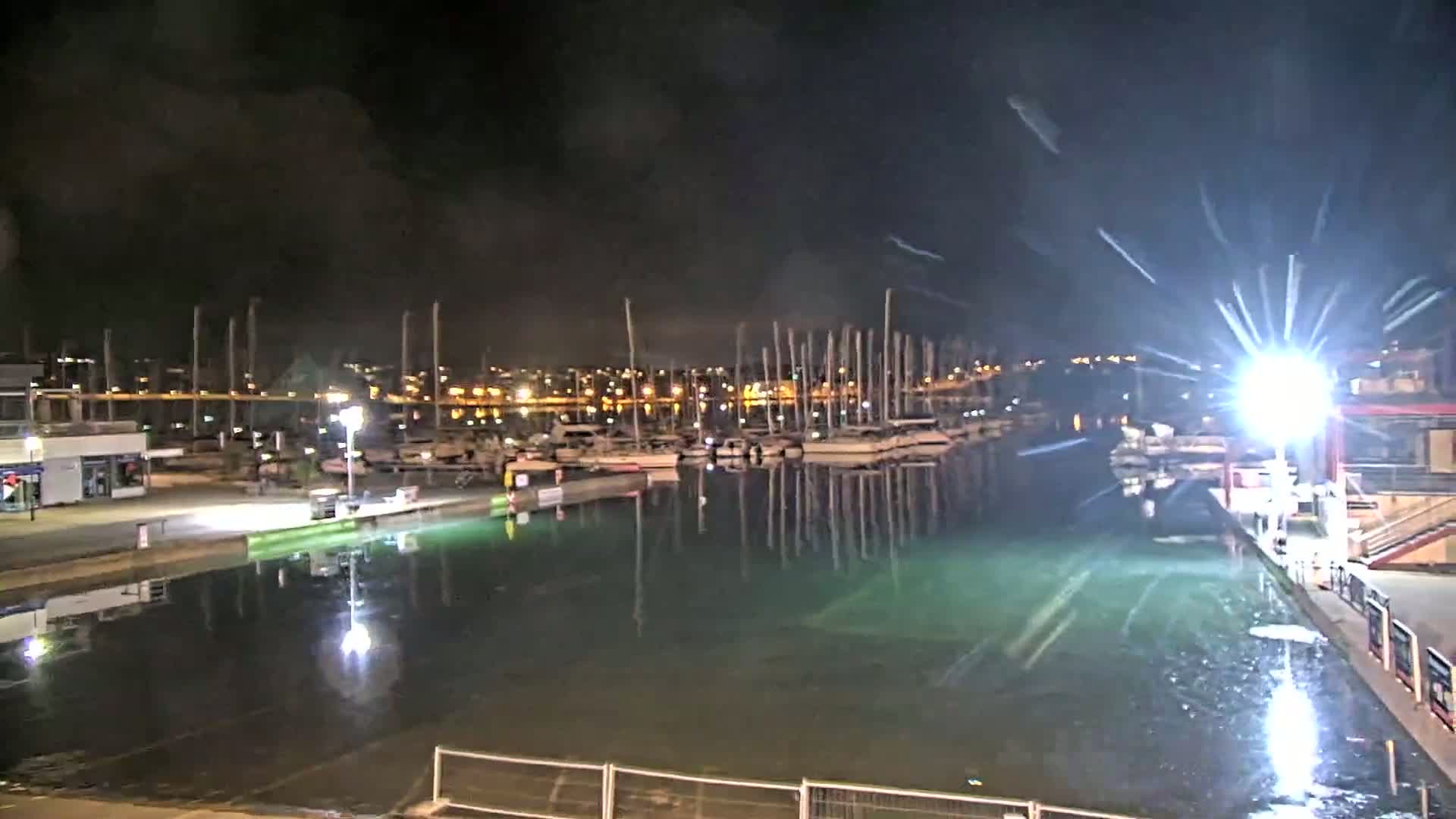 A nighttime view of a marina with numerous boats docked, reflecting in calm, greenish water under a dark, cloudy sky.