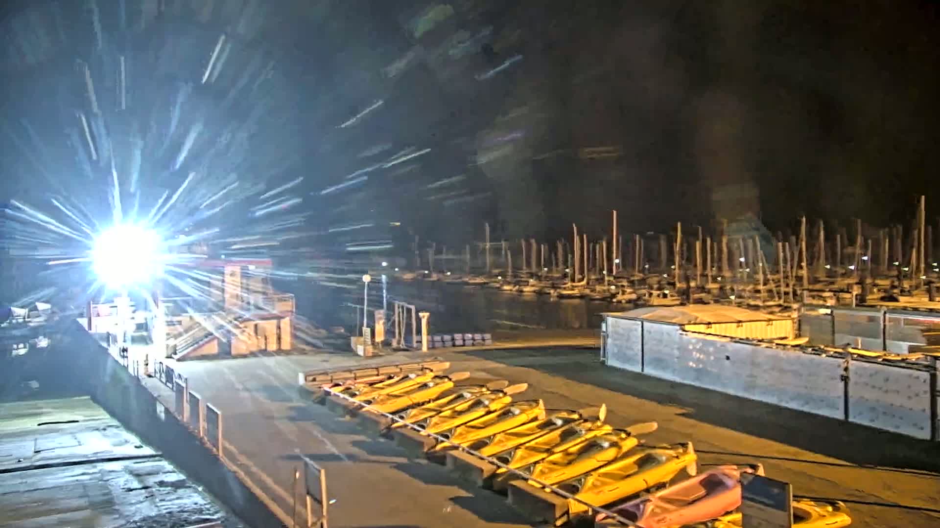 A brightly lit dock at night shows numerous yellow kayaks lined up, with a marina full of sailboats in the background.