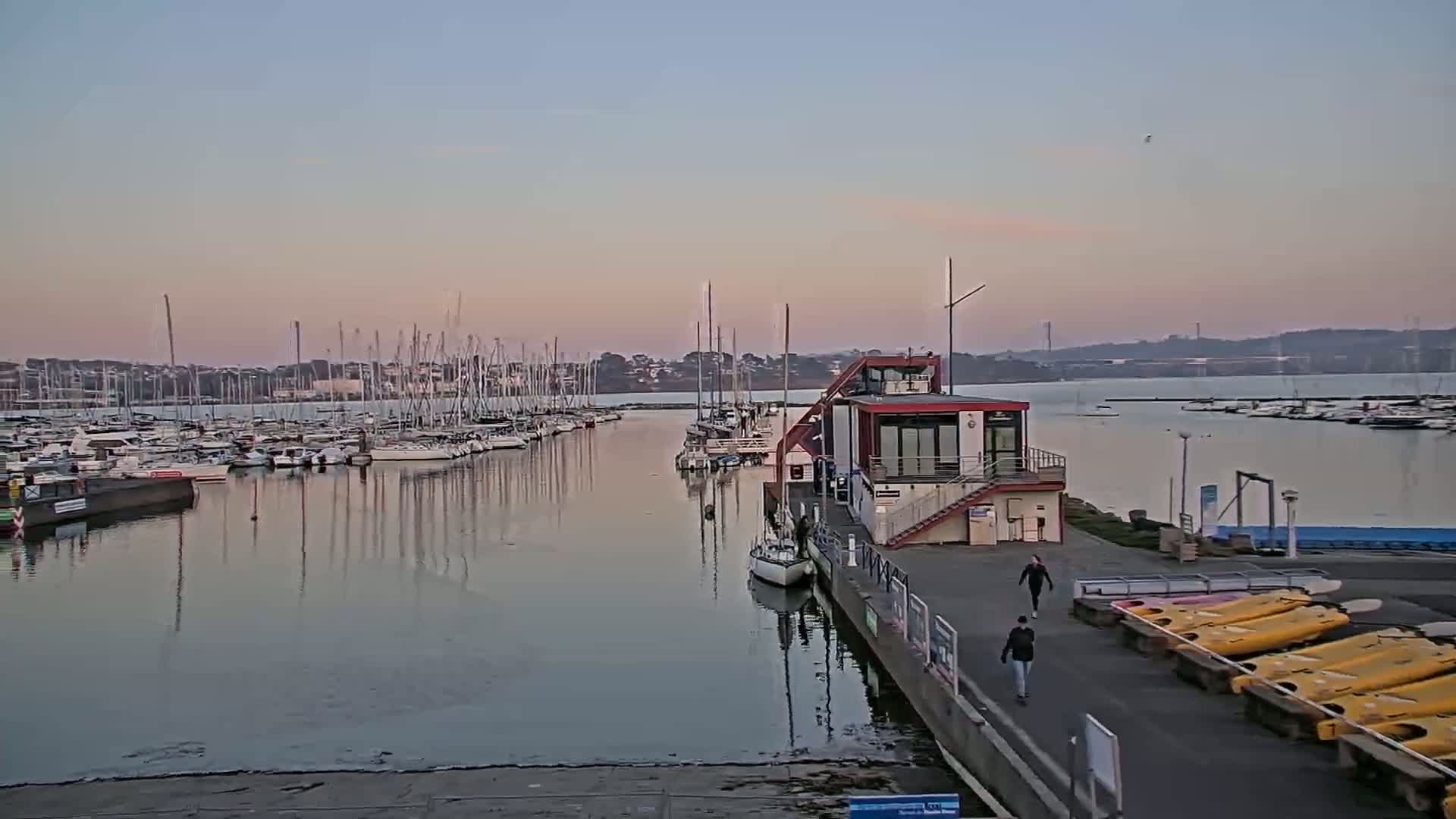 A calm marina at sunset, filled with numerous sailboats and small motorboats, features a modern building on a dock with a few people walking by.